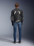 Vista posteriore della giacca in pelle Alpinestars GP Plus R V4 Rideknit, nero e bianco, con un grande logo bianco sulla parte bassa della schiena e branding sulle maniche, indossata con jeans da moto blu e stivali neri, evidenziando la costruzione tecnica e la vestibilità sportiva.