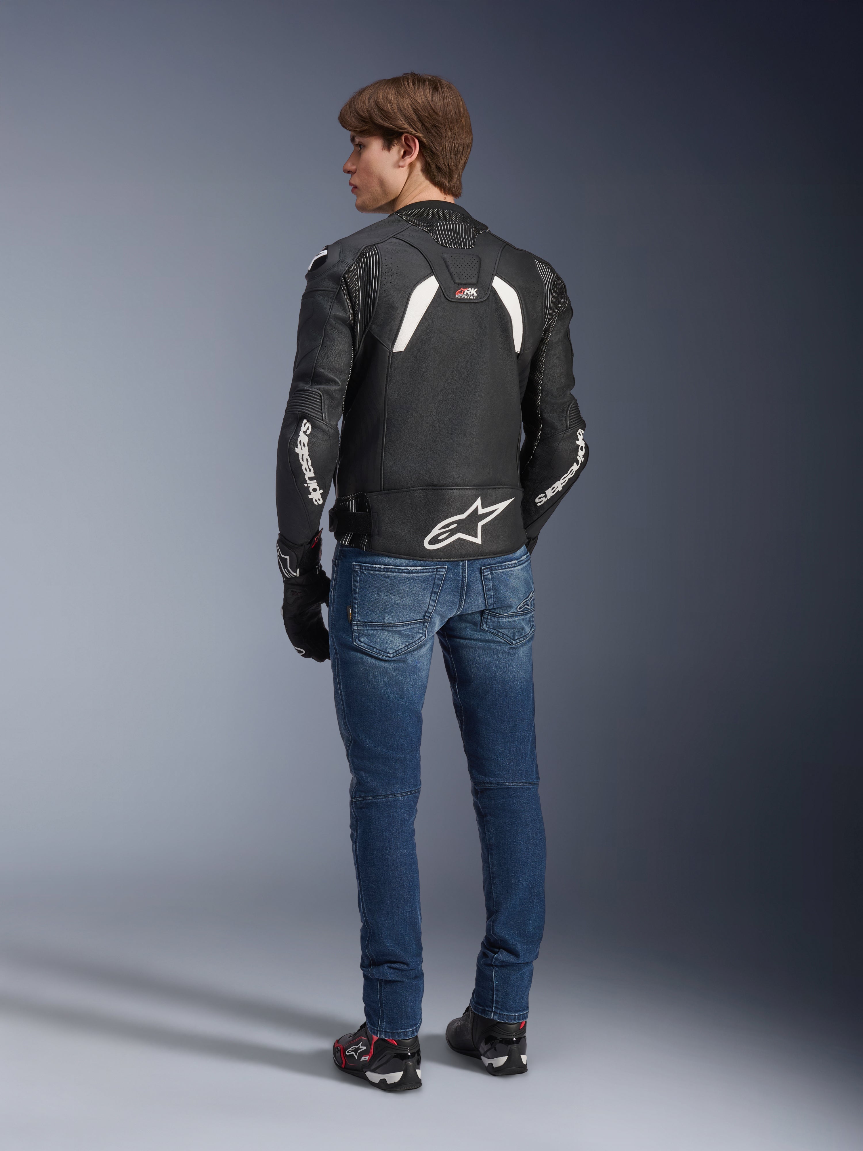 Giacca in pelle Alpinestars GP Plus R V4 Rideknit in nero e bianco, vista posteriore su un modello, con marchio Alpinestars bianco sulle maniche e un grande logo a stella sulla parte inferiore della schiena, indossata con jeans blu e scarpe da moto nere, abbigliamento tecnico da moto