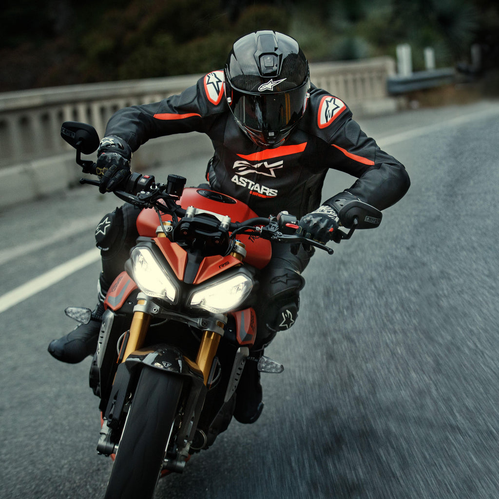 Motociclista alla guida di una moto sportiva rossa, giacca in pelle Alpinestars GP Plus R V4 Rideknit, nera con dettagli rosso fluo e bianchi, con logo Alpinestars bianco e scritta 'ASTARS', abbinata a casco racing e guanti neri, abbigliamento tecnico in azione su strada.