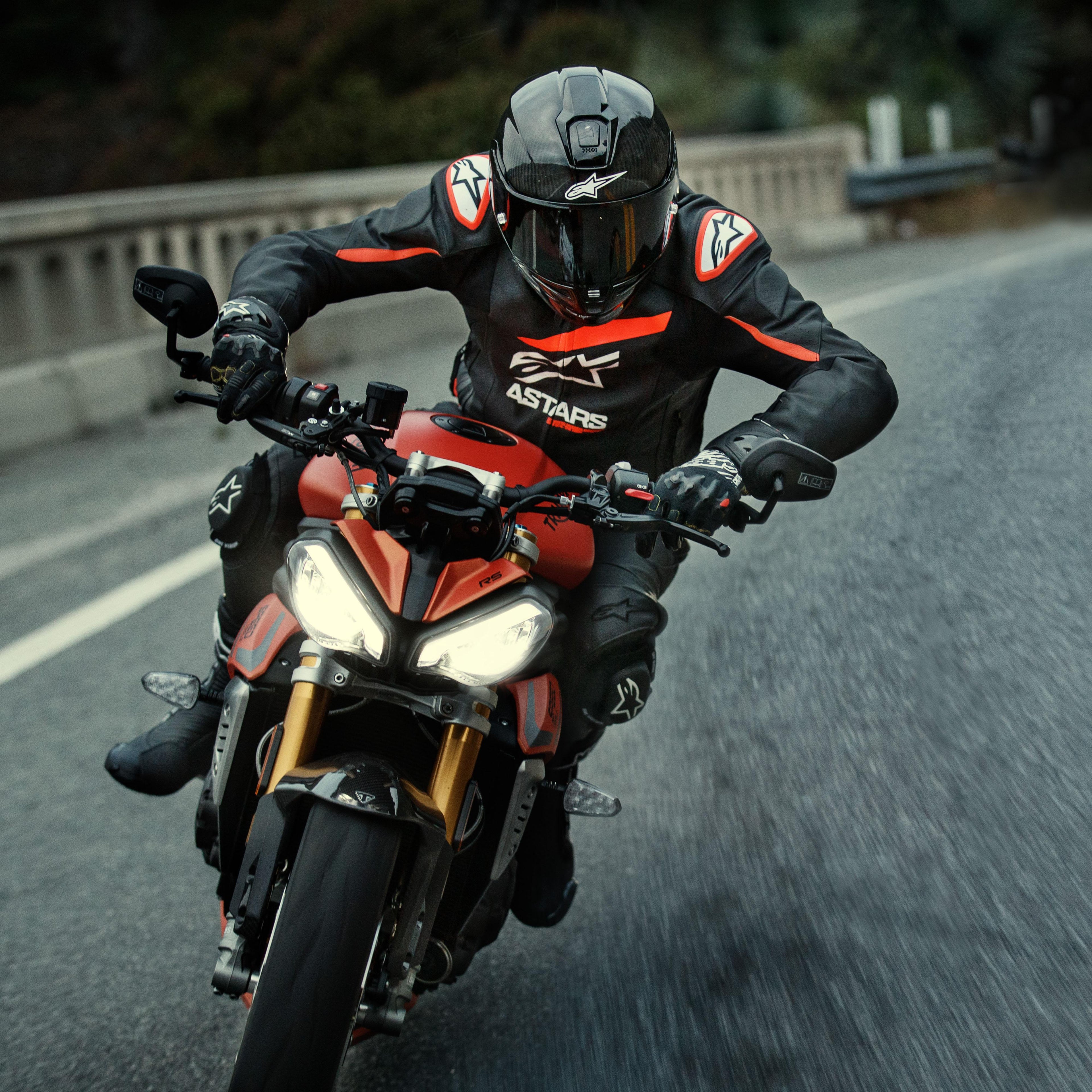 Motociclista alla guida di una moto sportiva rossa, giacca in pelle Alpinestars GP Plus R V4 Rideknit, nera con dettagli rosso fluo e bianchi, con logo Alpinestars bianco e scritta 'ASTARS', abbinata a casco racing e guanti neri, abbigliamento tecnico in azione su strada.