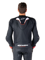 Vista posteriore di un pilota che indossa la giacca in pelle Alpinestars Missile V3 Ignition, in pelle nera con accenti bianchi e rossi, caratterizzata da una sottile gobba aerodinamica sportiva con logo bianco e marchio 'alpinestars RACING AHEAD' sulla parte bassa della schiena, abbinata a pantaloni da corsa e guanti in pelle nera coordinati.