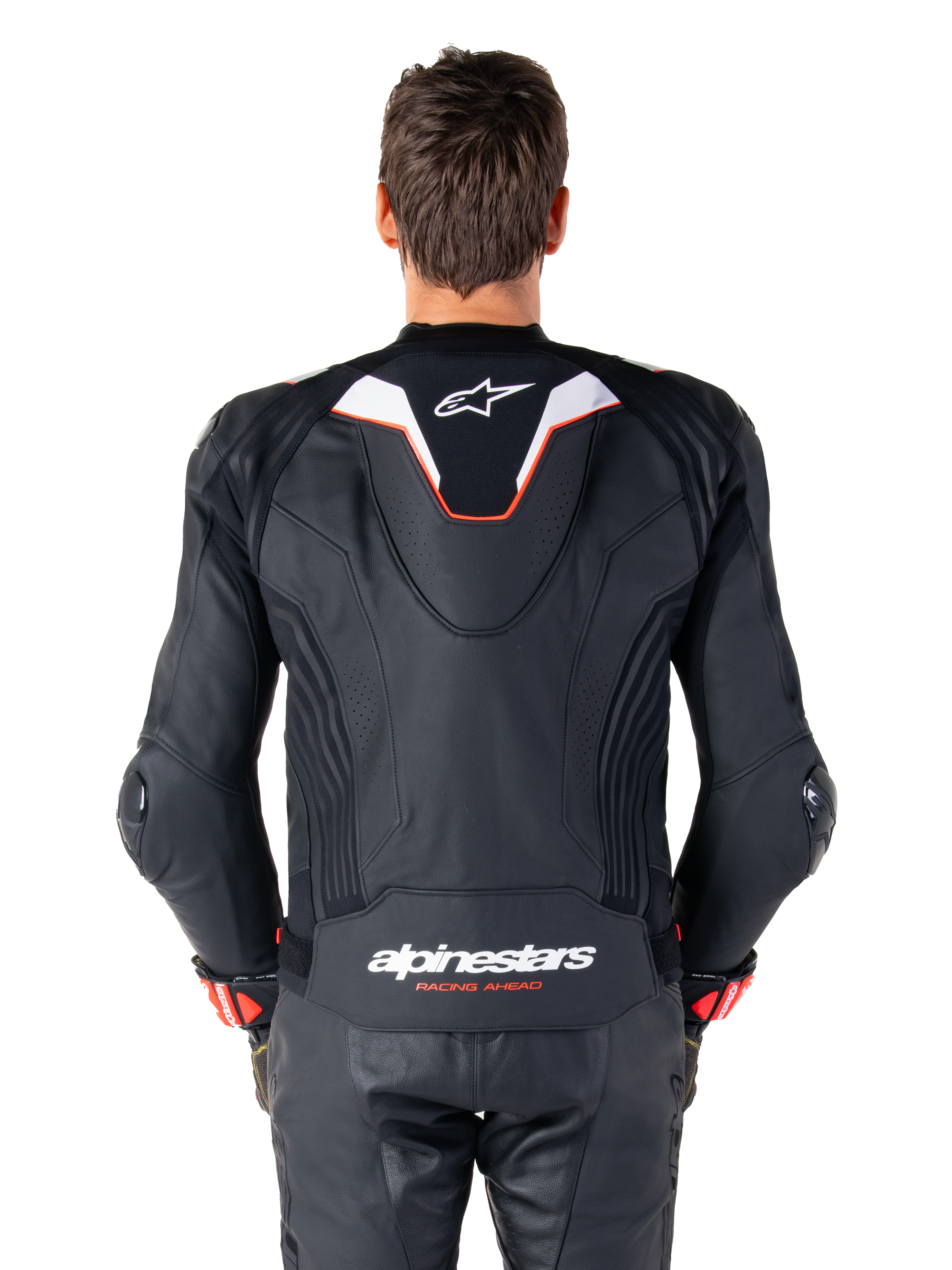 Vista posteriore di un pilota che indossa la giacca in pelle Alpinestars Missile V3 Ignition, in pelle nera con accenti bianchi e rossi, caratterizzata da una sottile gobba aerodinamica sportiva con logo bianco e marchio 'alpinestars RACING AHEAD' sulla parte bassa della schiena, abbinata a pantaloni da corsa e guanti in pelle nera coordinati.