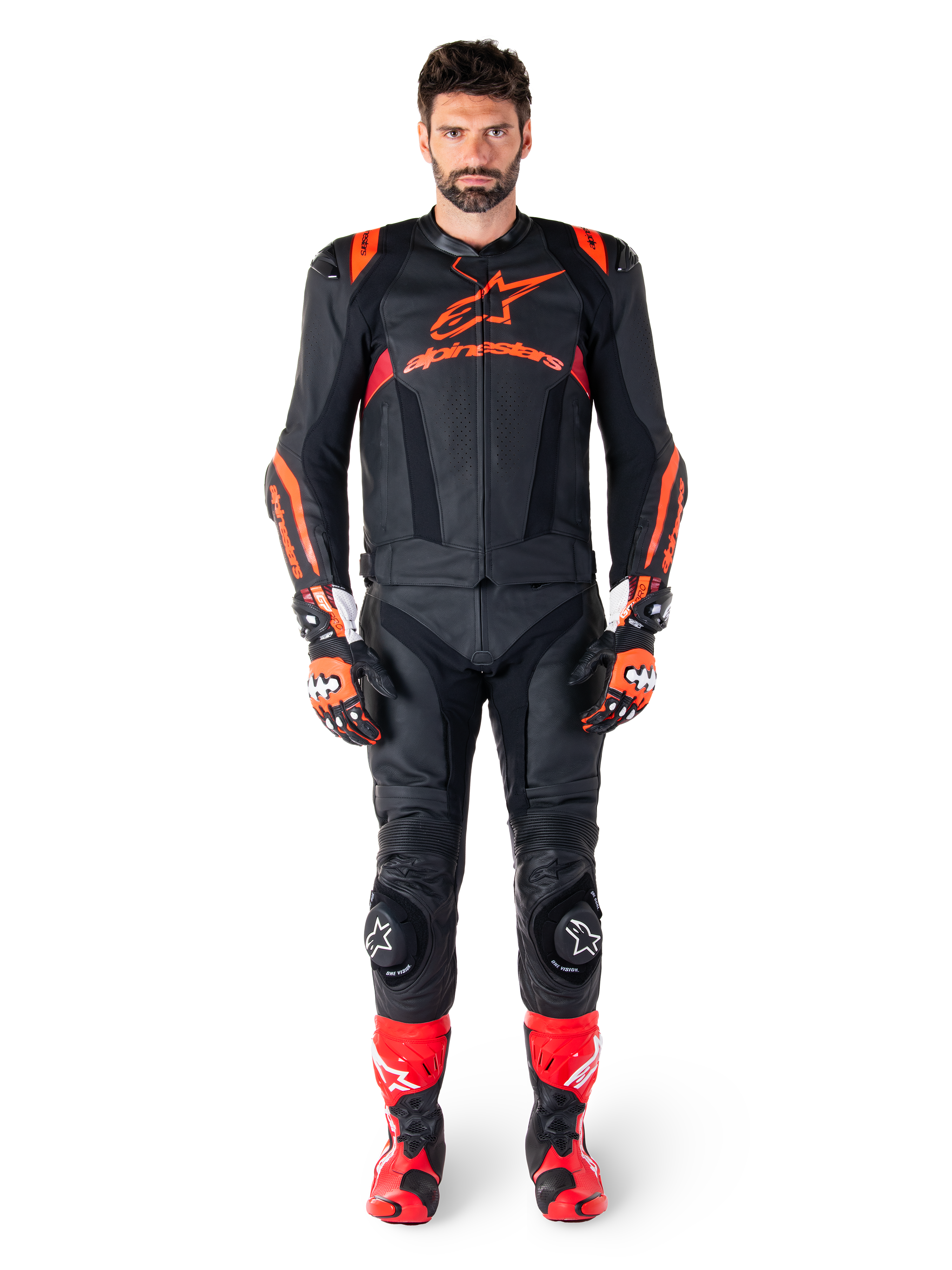 Modello maschile che indossa la giacca in pelle Alpinestars Missile V3 Ignition in Nero Rosso Fluo Rosso Scuro, equipaggiamento completo da gara motociclistica con pantaloni in pelle coordinati, guanti da corsa e stivali, caratterizzato dal logo Alpinestars sul petto, posa frontale, abbigliamento professionale per guida sportiva