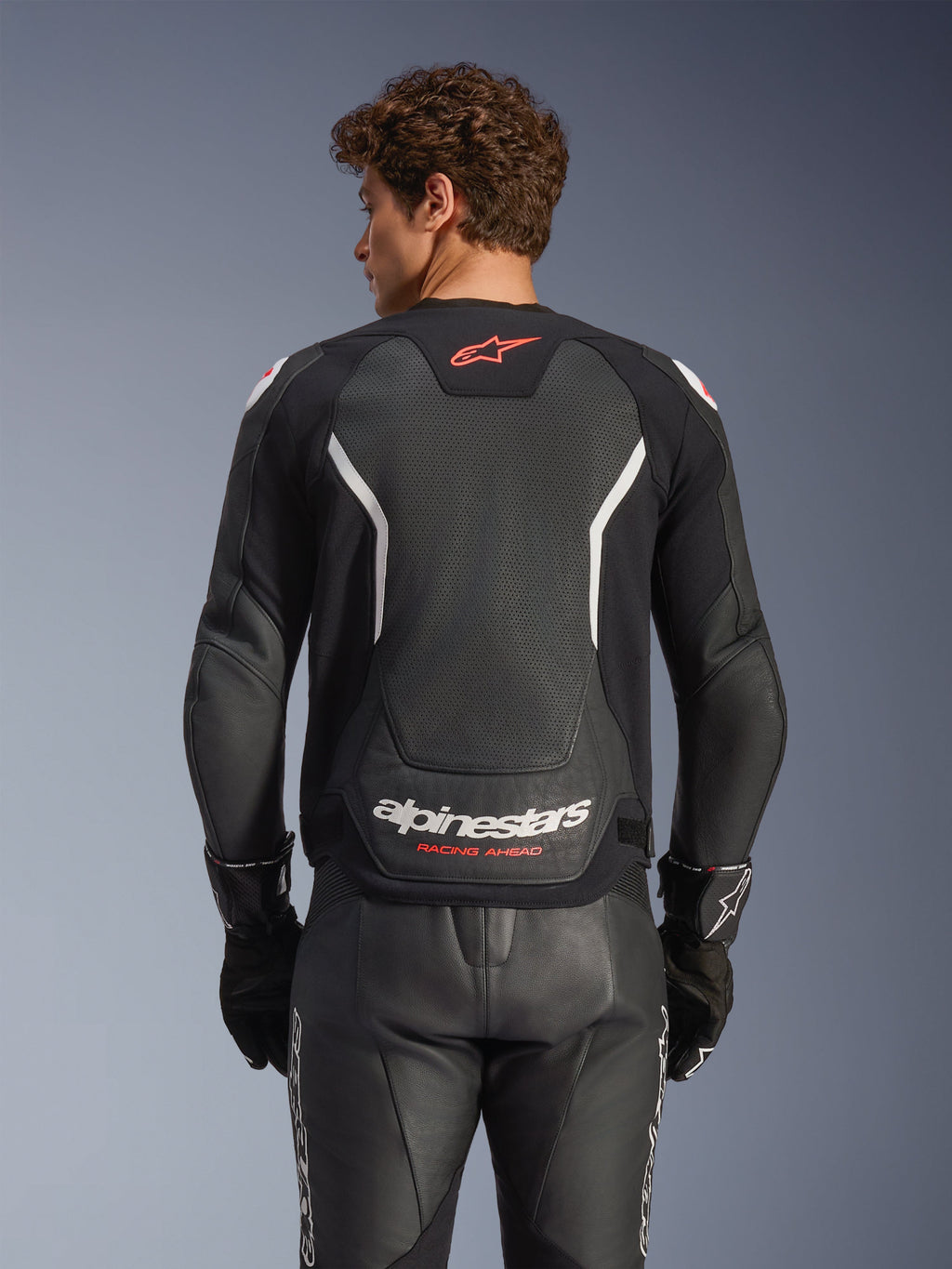 Giacca in pelle Alpinestars GP Force V2 Air, nero con accenti bianchi e rossi, vista posteriore con pannello posteriore in pelle traforata, branding Alpinestars bianco e dettagli del logo rossi, indossata con pantaloni racing e guanti in pelle nera coordinati.