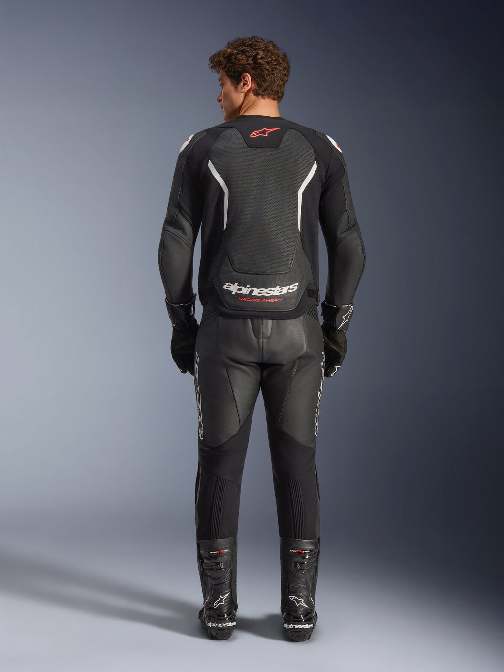 Giacca in pelle Alpinestars GP Force V2 Air in nero e bianco, vista posteriore, con ampie traforature per il flusso d'aria, dettagli bianchi sulle spalle e branding evidente, indossata con pantaloni e stivali da moto in pelle nera