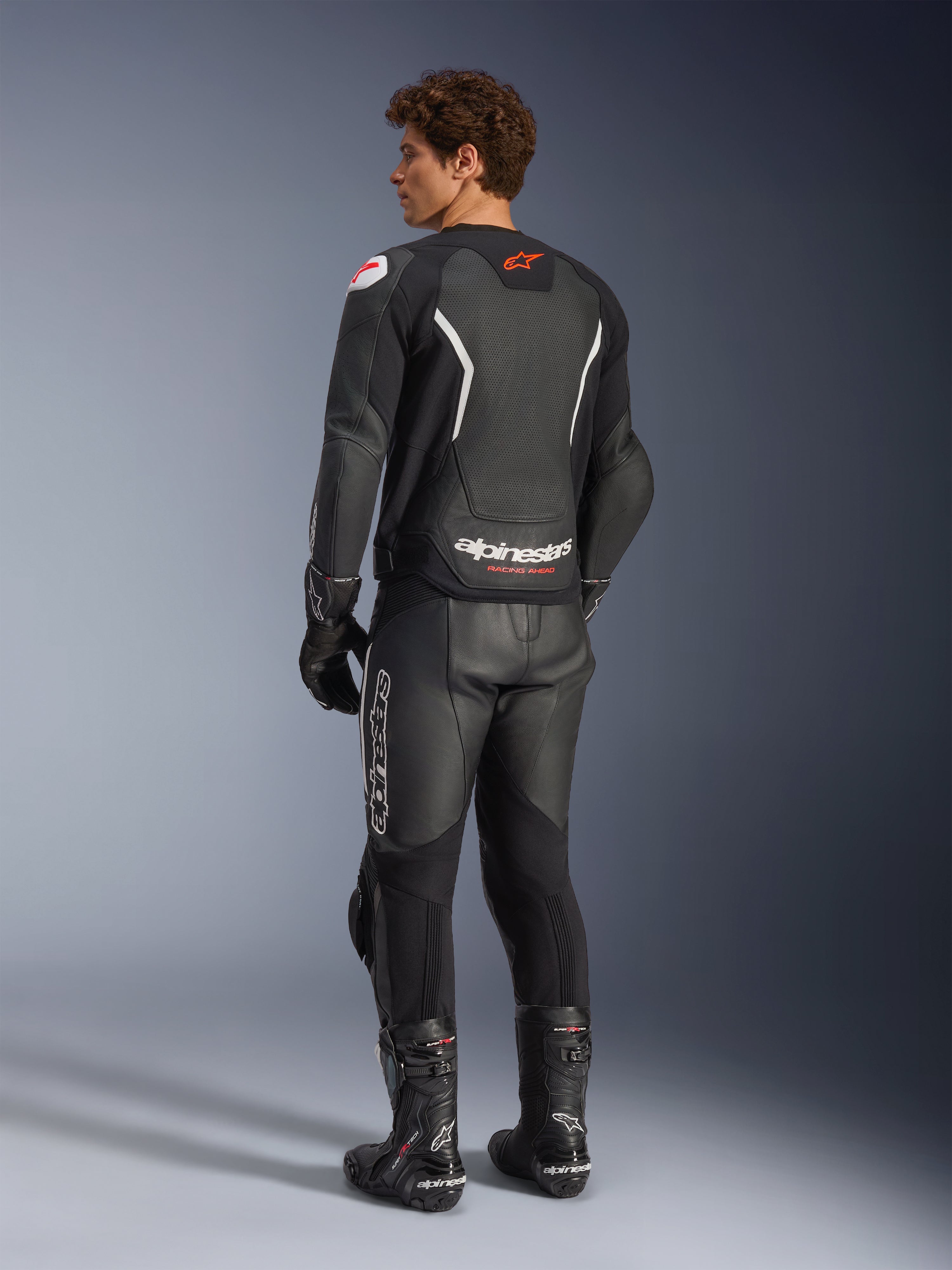 Giacca in pelle Alpinestars GP Force V2 Air, nero e bianco, vista posteriore su modello con pannello posteriore traforato per la ventilazione, branding bianco e logo a stella rosso, indossata con pantaloni da corsa in pelle abbinati, guanti e stivali