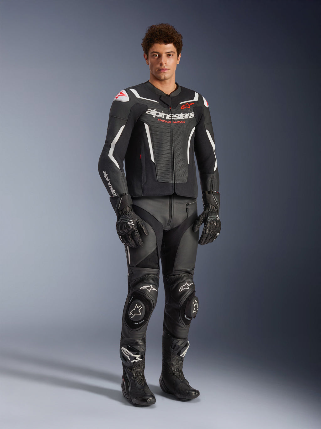Giacca in pelle Alpinestars GP Force V2 Air in nero e bianco, indossata da un pilota uomo con pantaloni racing, guanti e stivali neri coordinati, con pannelli in pelle traforata, accenti bianchi e logo Alpinestars rosso sul petto, abbigliamento sportivo da moto
