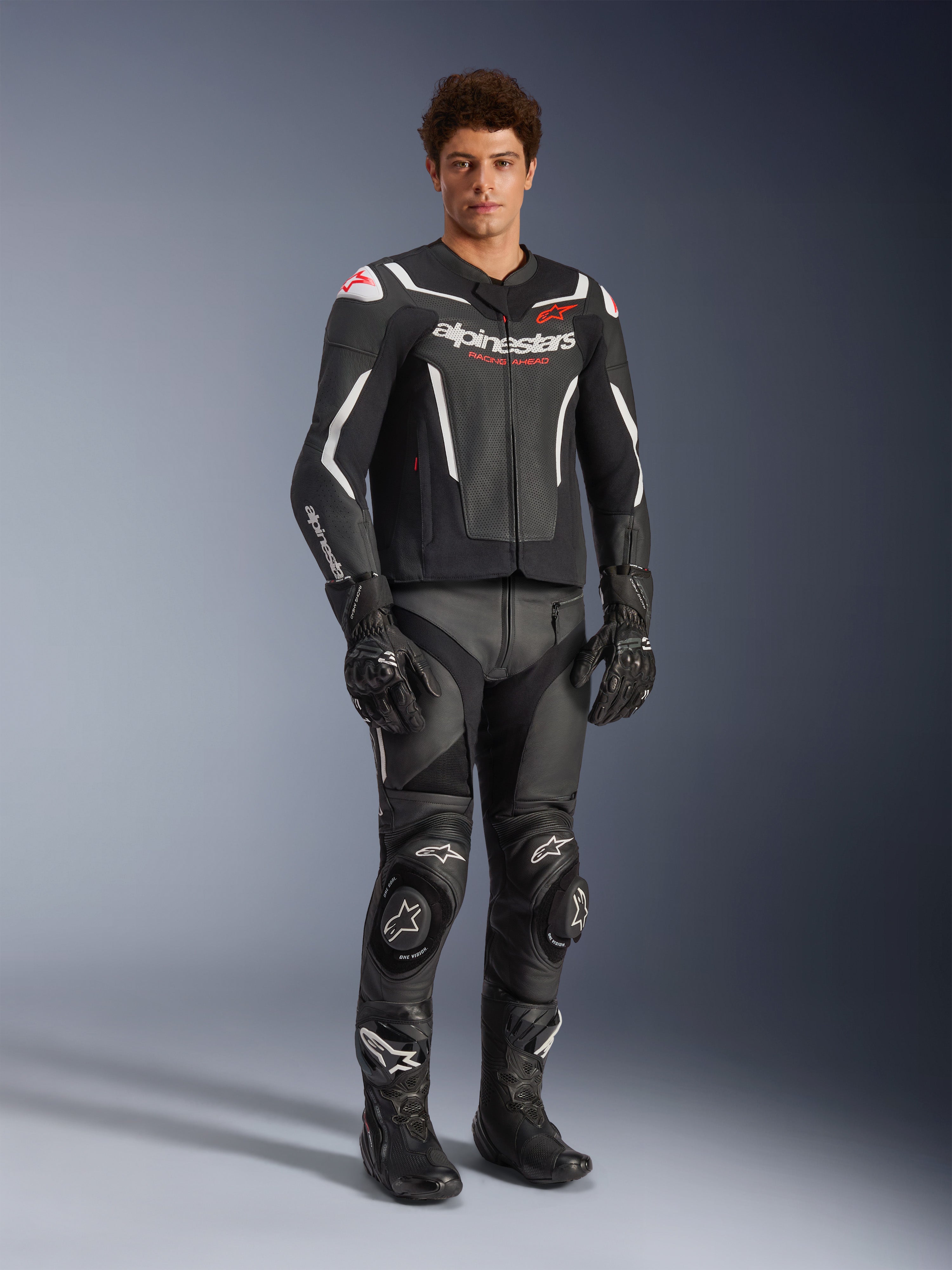Giacca in pelle Alpinestars GP Force V2 Air in nero e bianco, indossata da un pilota uomo con pantaloni racing, guanti e stivali neri coordinati, con pannelli in pelle traforata, accenti bianchi e logo Alpinestars rosso sul petto, abbigliamento sportivo da moto