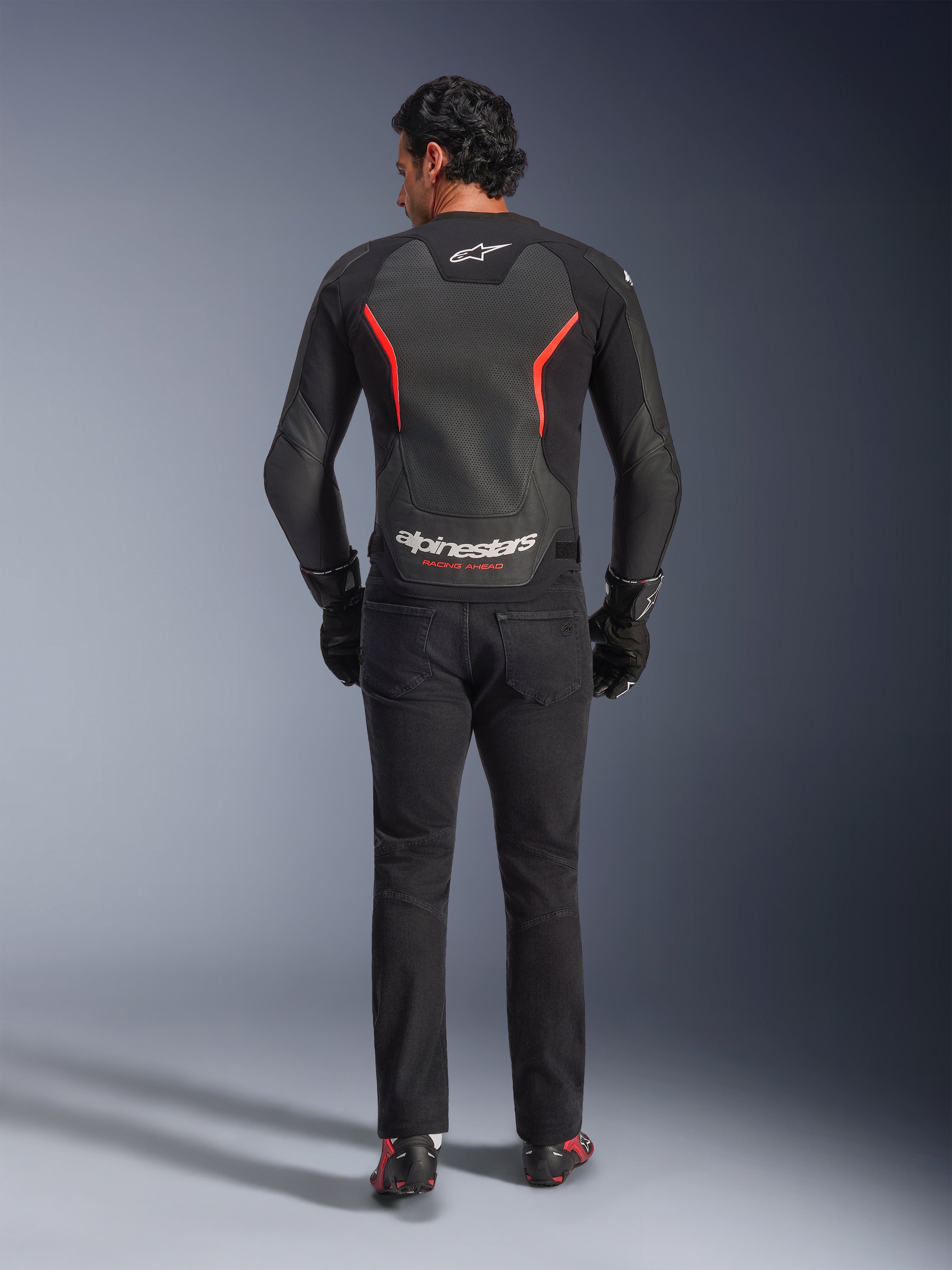 Giacca in pelle Alpinestars GP Force V2 Air, nero e rosso fluo, vista posteriore su modello, con pannelli in pelle traforata sul retro, accenti rossi e logo Alpinestars, abbinata a jeans da moto neri e guanti per un look sportivo.