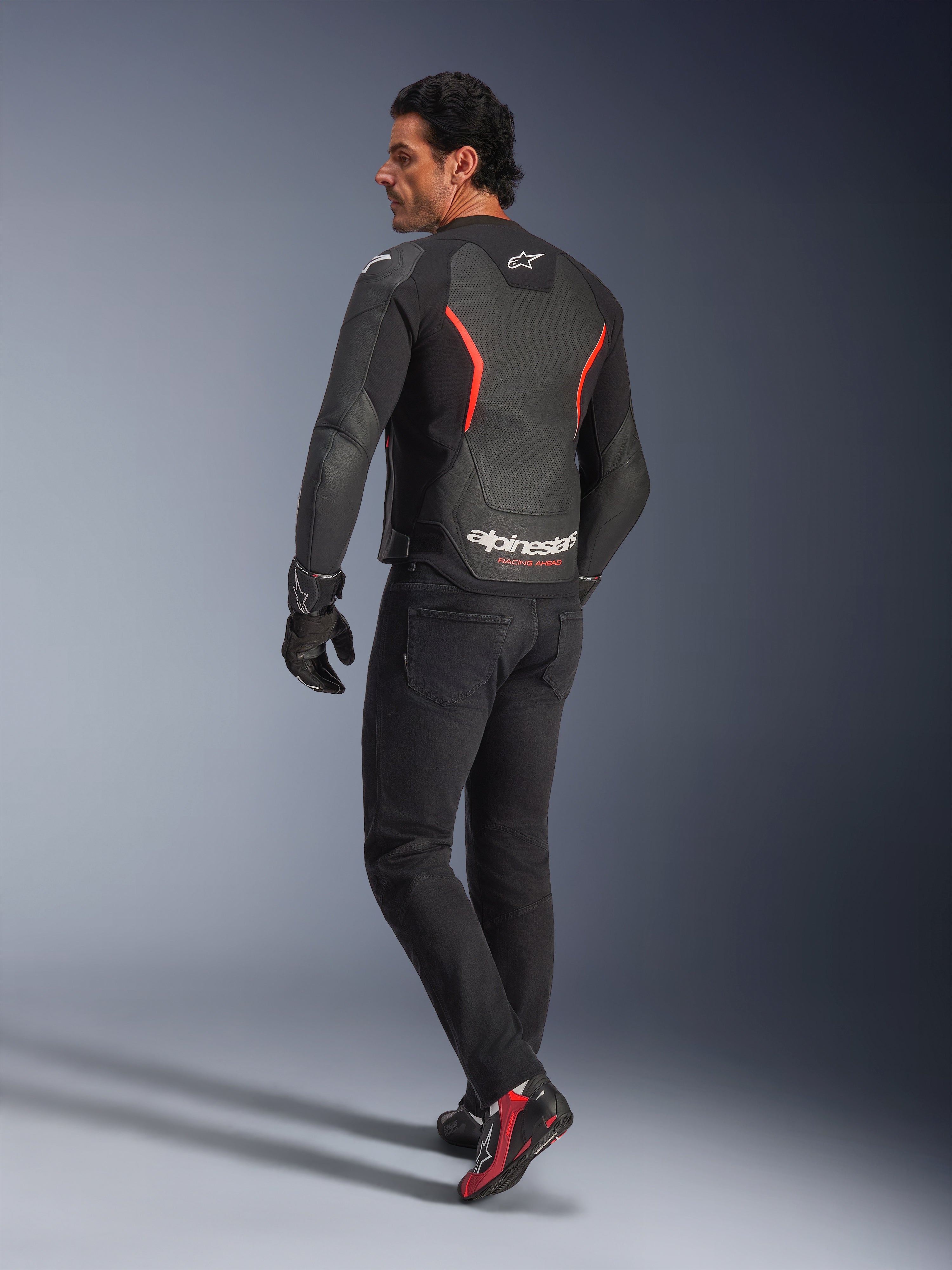 Modello visto da dietro che indossa la giacca in pelle Alpinestars GP Force V2 Air in Black Red Fluo, con ampie perforazioni sulla schiena e logo bianco, abbinata a jeans da moto neri, guanti e stivali da moto neri e rossi in un contesto da studio.