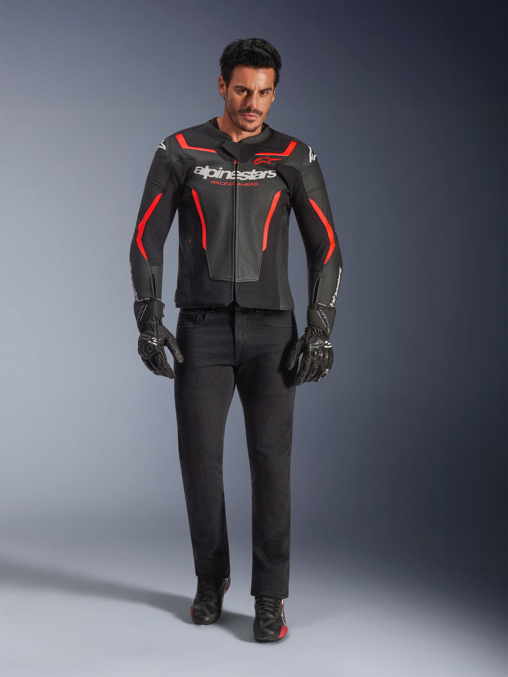 Pilota con giacca in pelle Alpinestars GP Force V2 Air, nero e rosso fluo, abbinata a jeans da moto neri, guanti e scarpe, con pelle traforata, logo bianco e accenti rossi per un look sportivo