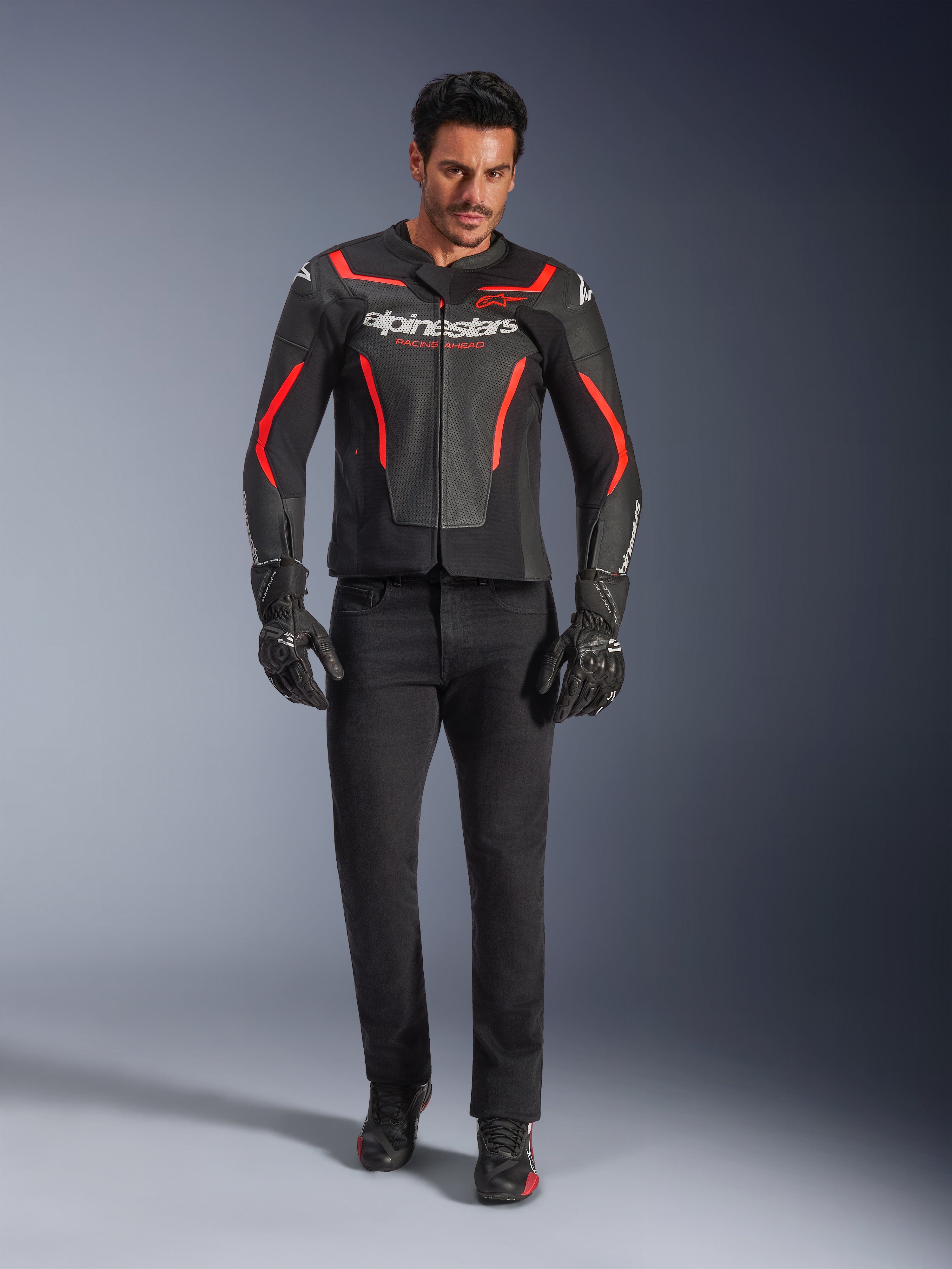 Pilota con giacca in pelle Alpinestars GP Force V2 Air, nero e rosso fluo, abbinata a jeans da moto neri, guanti e scarpe, con pelle traforata, logo bianco e accenti rossi per un look sportivo