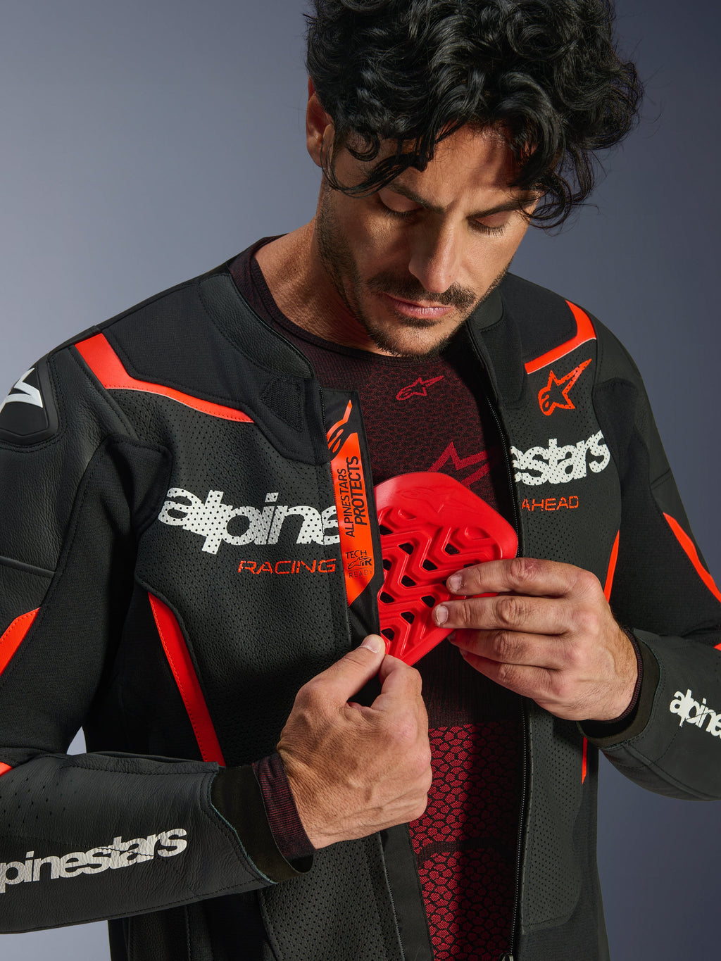 Giacca in pelle Alpinestars GP Force V2 Air in nero e rosso fluo, uomo che apre la zip della giacca per mostrare l'inserimento di una protezione per il petto rossa, caratterizzata da pelle traforata, logo bianco evidente e dettaglio Tech-Air Ready sulla patta interna
