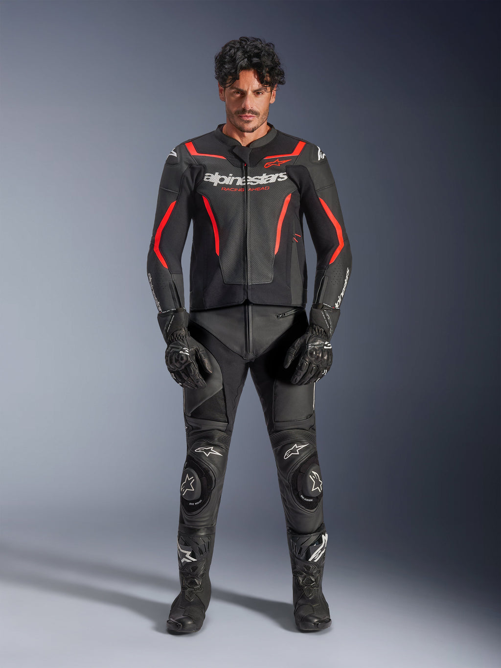 Modello che indossa la giacca in pelle Alpinestars GP Force V2 Air in nero e rosso fluo, con pannelli traforati sul petto e loghi bianchi, abbinata a pantaloni in pelle da moto neri, guanti neri e stivali neri per un look racing completo.