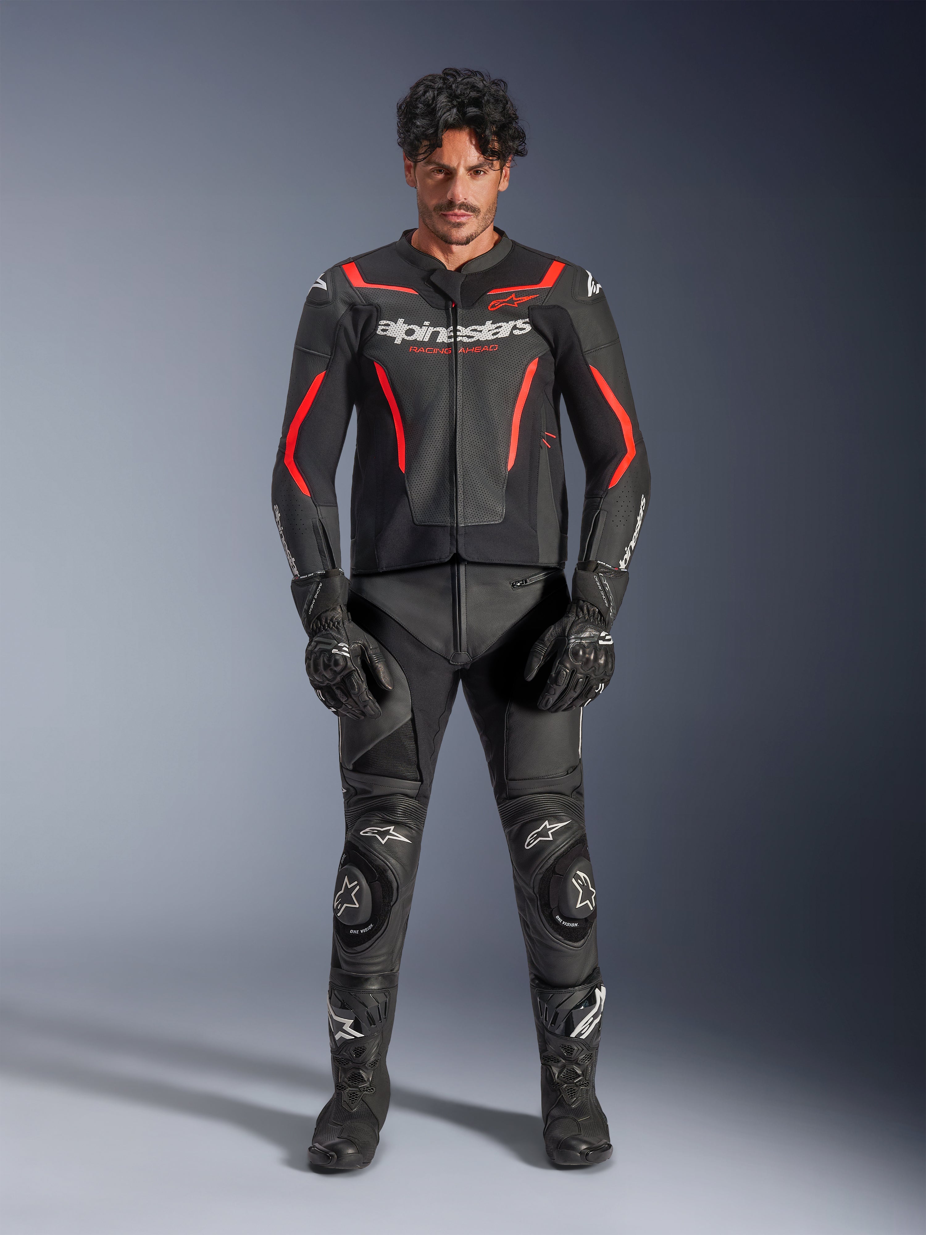 Modello che indossa la giacca in pelle Alpinestars GP Force V2 Air in nero e rosso fluo, con pannelli traforati sul petto e loghi bianchi, abbinata a pantaloni in pelle da moto neri, guanti neri e stivali neri per un look racing completo.