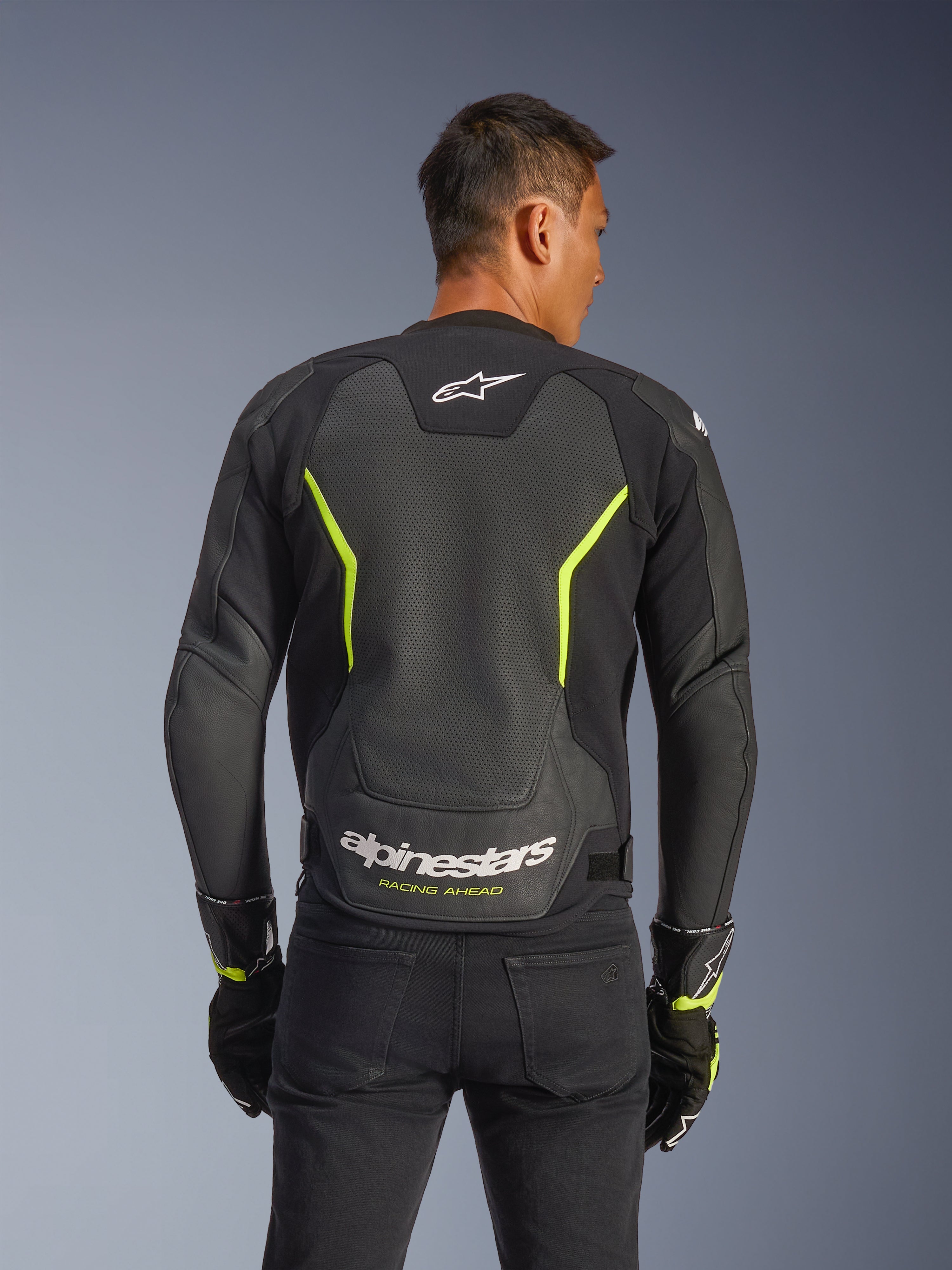Giacca in pelle Alpinestars GP Force V2 Air, nero e giallo fluo, vista posteriore di un motociclista che mostra ampi pannelli in pelle traforata per la circolazione dell'aria, branding Alpinestars bianco e scritta "Racing Ahead" gialla sulla parte inferiore della schiena, abbigliamento sportivo da moto per guida su strada.
