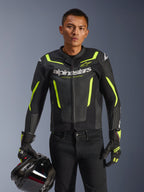 Pilota uomo che indossa la giacca in pelle Alpinestars GP Force V2 Air in nero e giallo fluo, tenendo un casco da moto nero, caratterizzata da pelle traforata con vistoso branding Alpinestars bianco e accenti giallo neon, abbinata a guanti da corsa coordinati e pantaloni neri