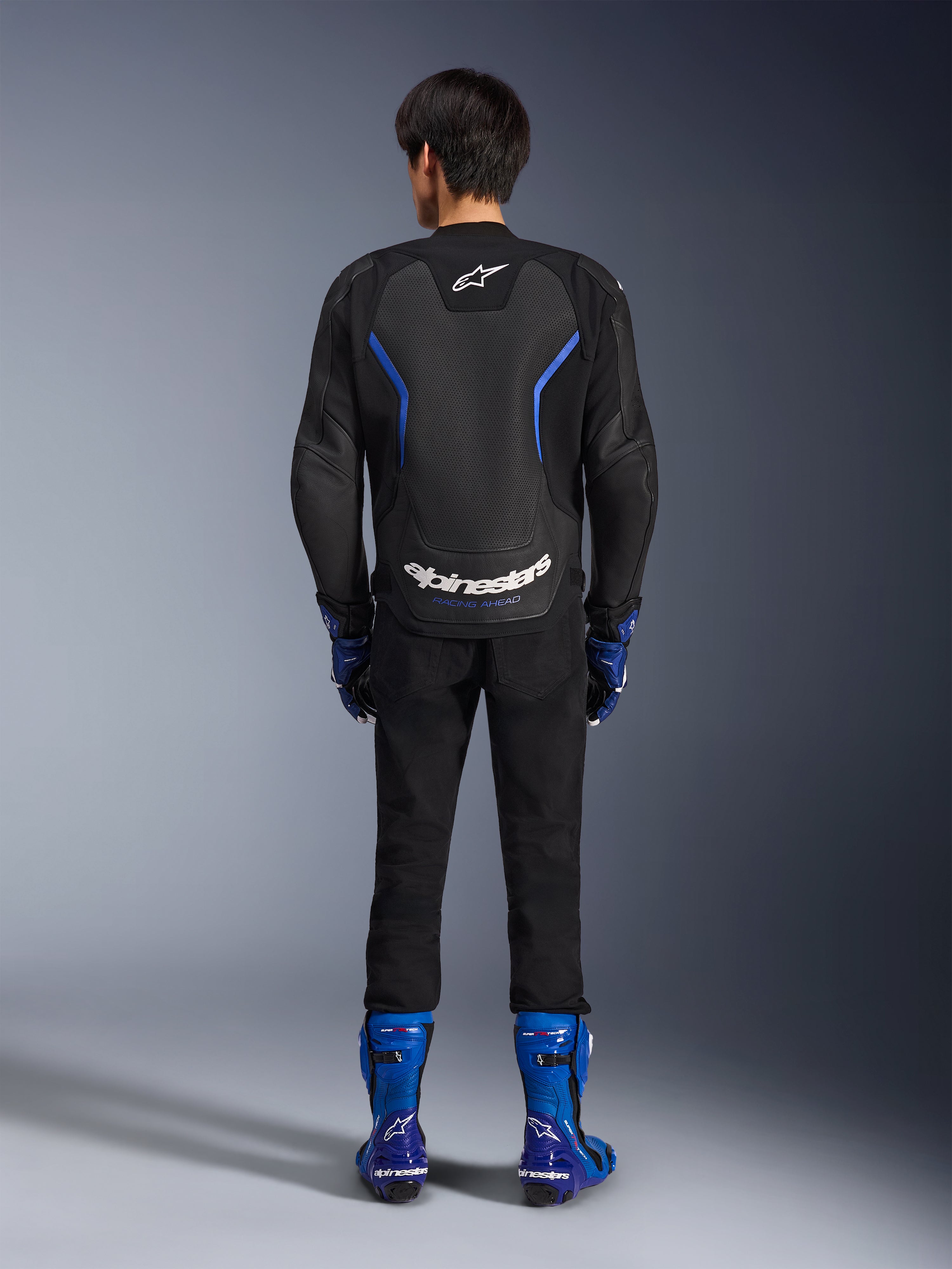 Giacca in pelle Alpinestars GP Force V2 Airflow, Black Metallic Blue, vista posteriore con pannelli in pelle traforata e logo bianco, indossata con stivali racing blu e pantaloni neri, abbigliamento sportivo da moto racing