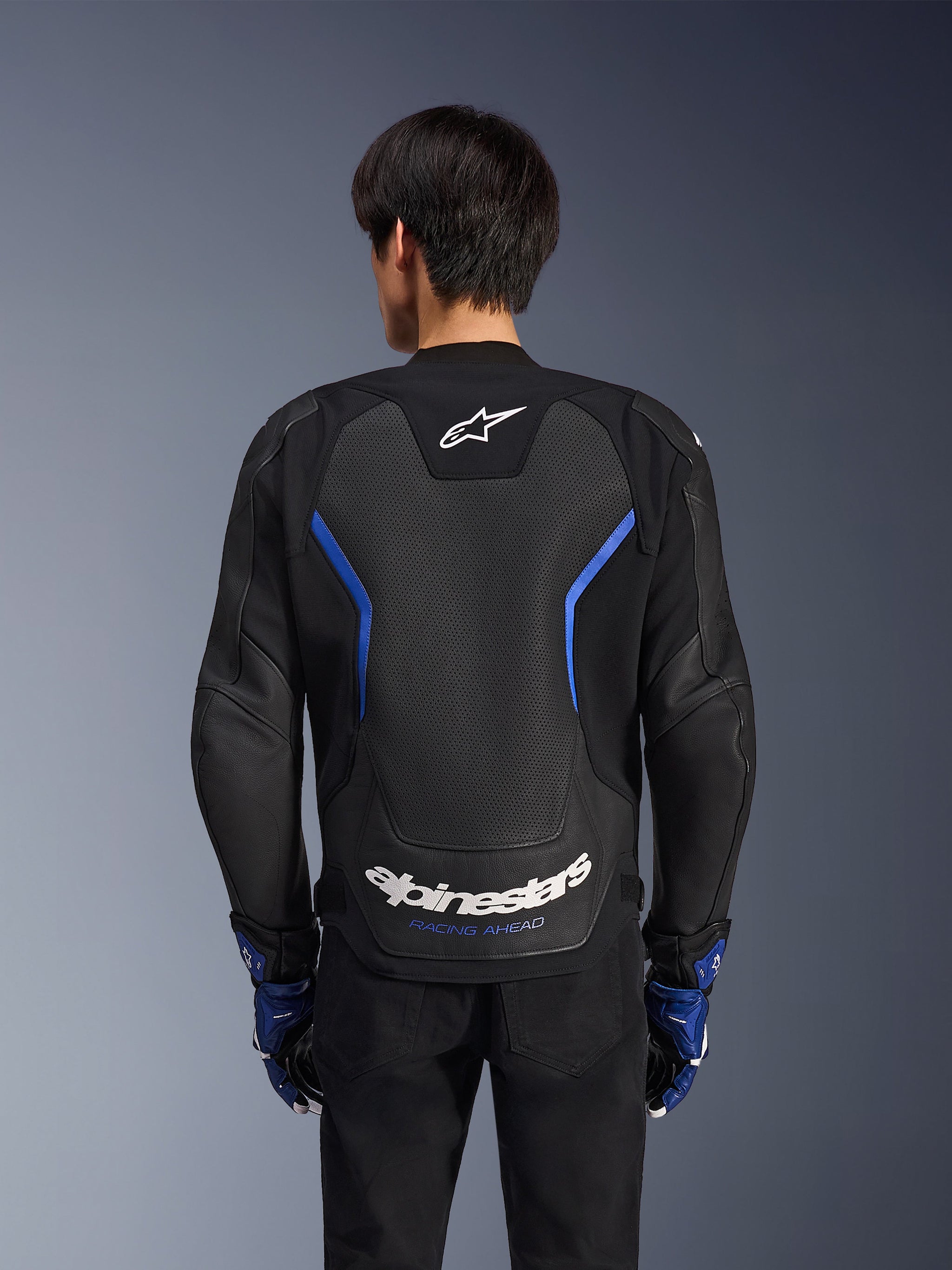 Alpinestars GP Force V2 Airflow Leather Jacket, giacca in pelle, nero blu metallizzato, vista posteriore con ampio pannello traforato per la ventilazione, accenti blu e marchio Alpinestars bianco, indossata con guanti blu e pantaloni neri per guida sportiva.