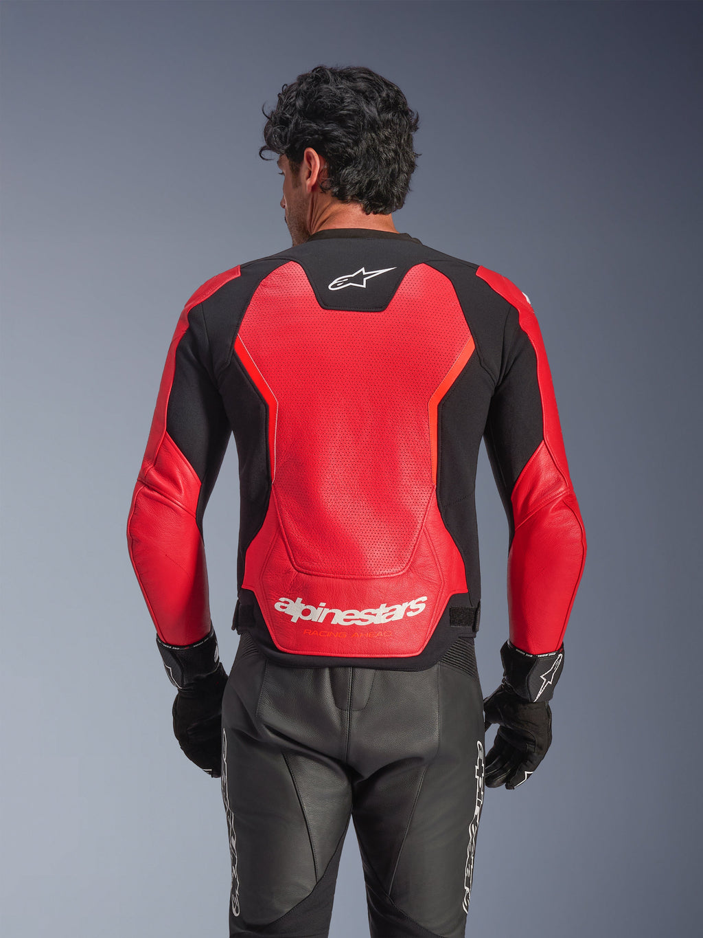 Giacca in pelle Alpinestars GP Force V2 Air in rosso e nero, vista posteriore che mostra la struttura in pelle traforata per la ventilazione, marchio Alpinestars bianco sulla parte inferiore della schiena e pannelli elasticizzati integrati, indossata con pantaloni e guanti da moto in pelle nera.