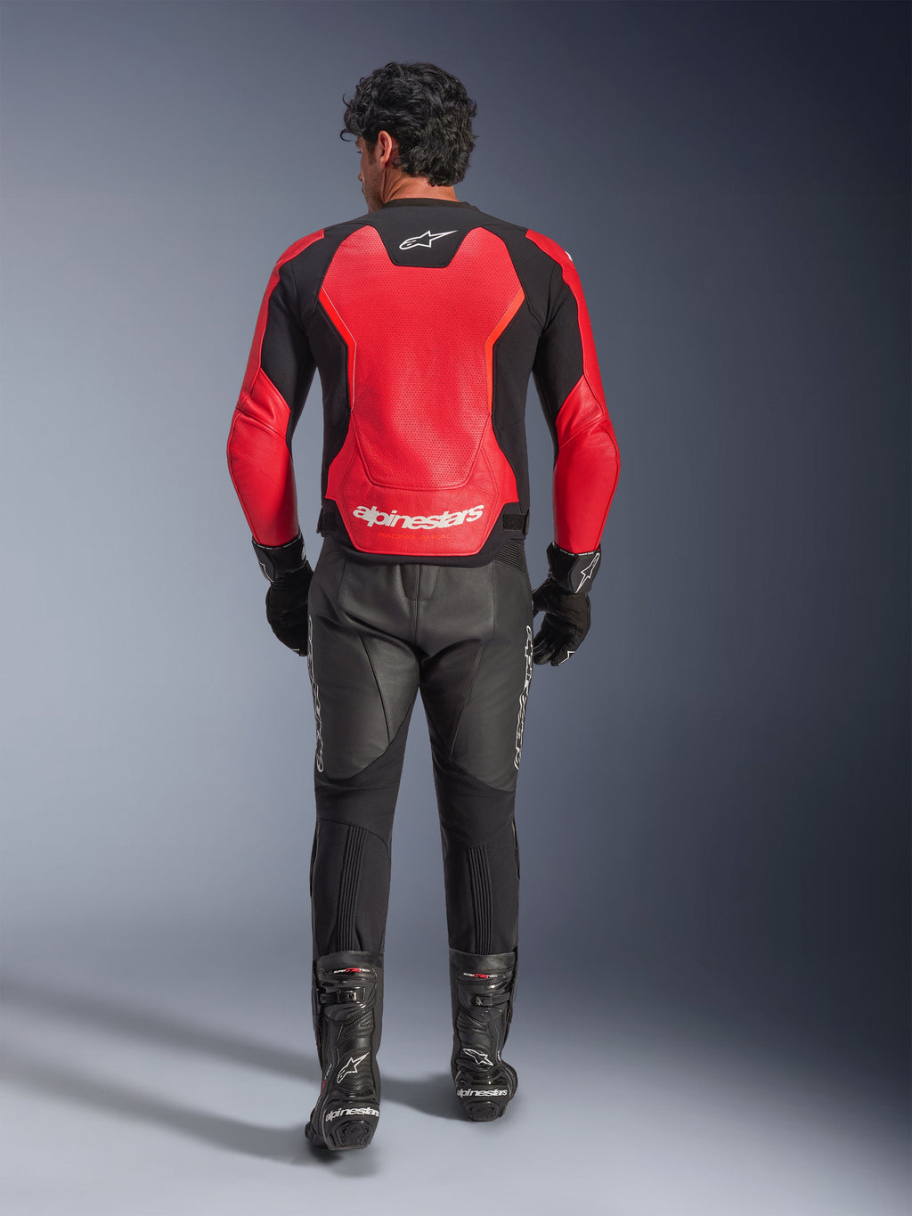 Vista posteriore di un pilota che indossa la giacca in pelle Alpinestars GP Force V2 Air in rosso e nero, con ampi pannelli traforati e logo Alpinestars bianco sul retro, abbinata a pantaloni da moto in pelle neri e stivali da corsa.