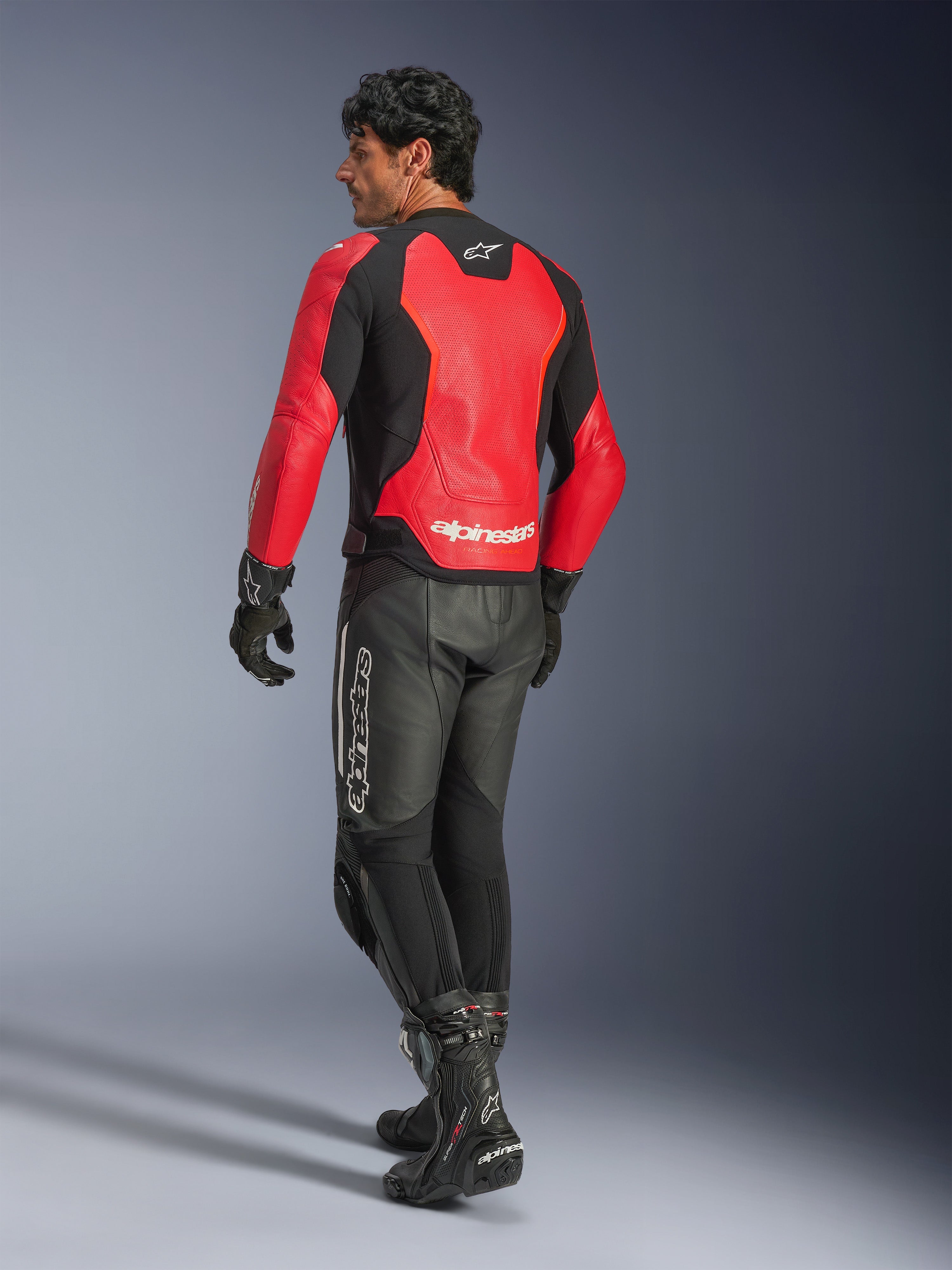 Giacca in pelle Alpinestars GP Force V2 Air, rossa e nera, vista posteriore su un pilota, indossata con pantaloni in pelle neri e stivali da corsa, con pannelli posteriori traforati e marchio Alpinestars