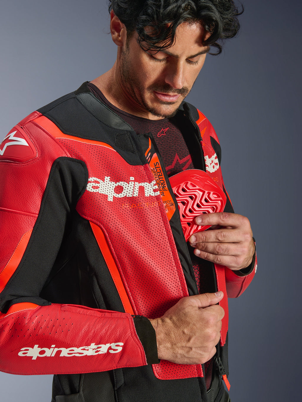 Giacca in pelle Alpinestars GP Force V2 Air, rosso e rosso fluo, primo piano di un pilota che inserisce una protezione pettorale rossa nella tasca interna, con pelle traforata e logo Tech-Air Ready, protezione premium per moto sportiva.