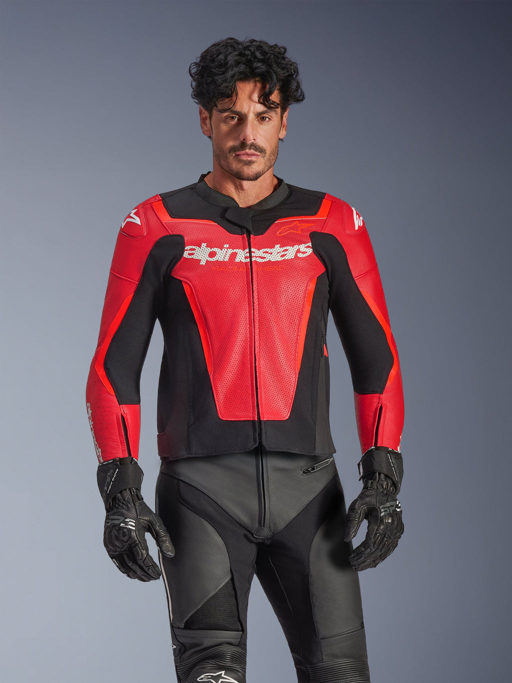 Alpinestars GP Force V2 Air Leather Jacket, giacca da moto racing rossa e nera, costruzione in pelle traforata con logo Alpinestars bianco, indossata da un modello e abbinata a pantaloni e guanti da corsa neri