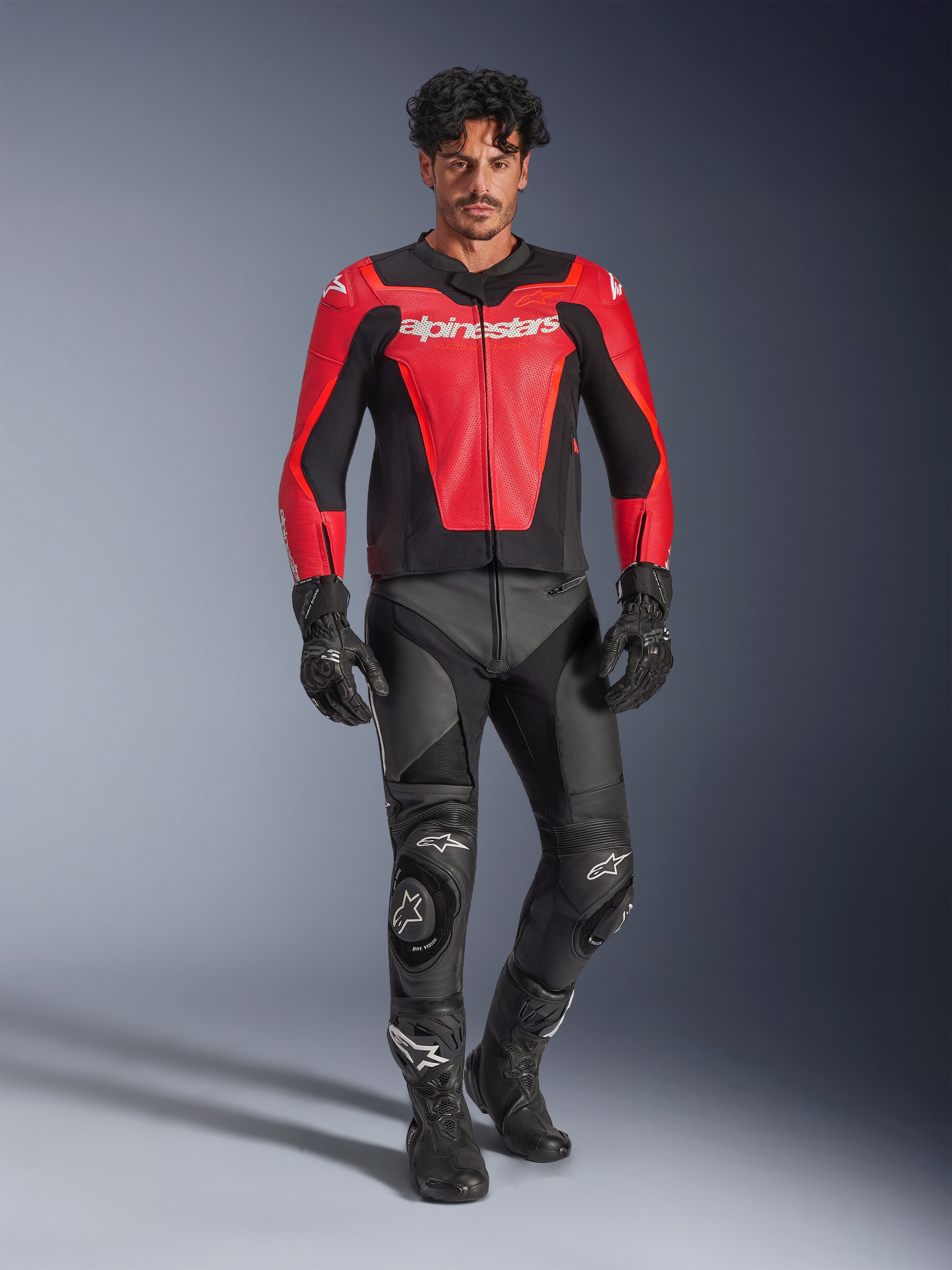 Giacca in pelle Alpinestars GP Force V2 Air, rosso e nero, pelle traforata con loghi bianchi, indossata da un modello con pantaloni racing e guanti in pelle nera, abbigliamento professionale da moto