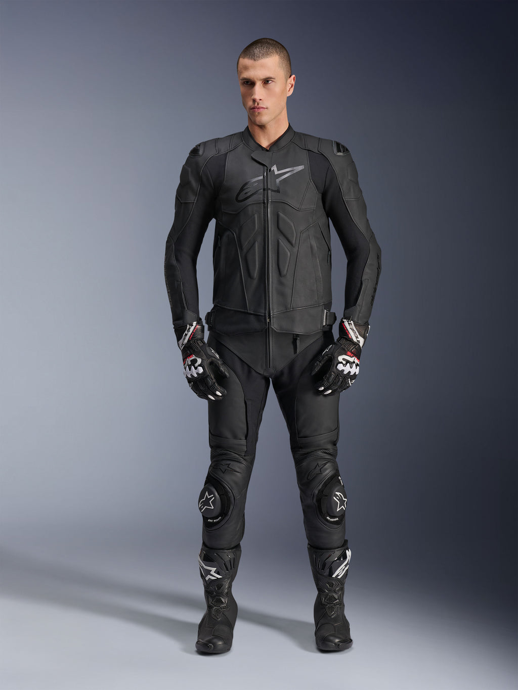 Giacca in pelle Alpinestars Dusk, nera, indossata da un modello uomo come parte di un outfit completo d'ispirazione racing con pantaloni in pelle abbinati, guanti e stivali, design ergonomico con protezioni sulle spalle e logo sul petto.
