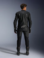 Pilota di moto in vista posteriore con giacca in pelle Alpinestars Dusk e pantaloni in pelle Missile V3, tuta in pelle nera con accenti bianchi, logo Alpinestars e marchio 'Dusk' sul retro, stivali e guanti racing neri, completo motorsport coordinato per corse su strada.