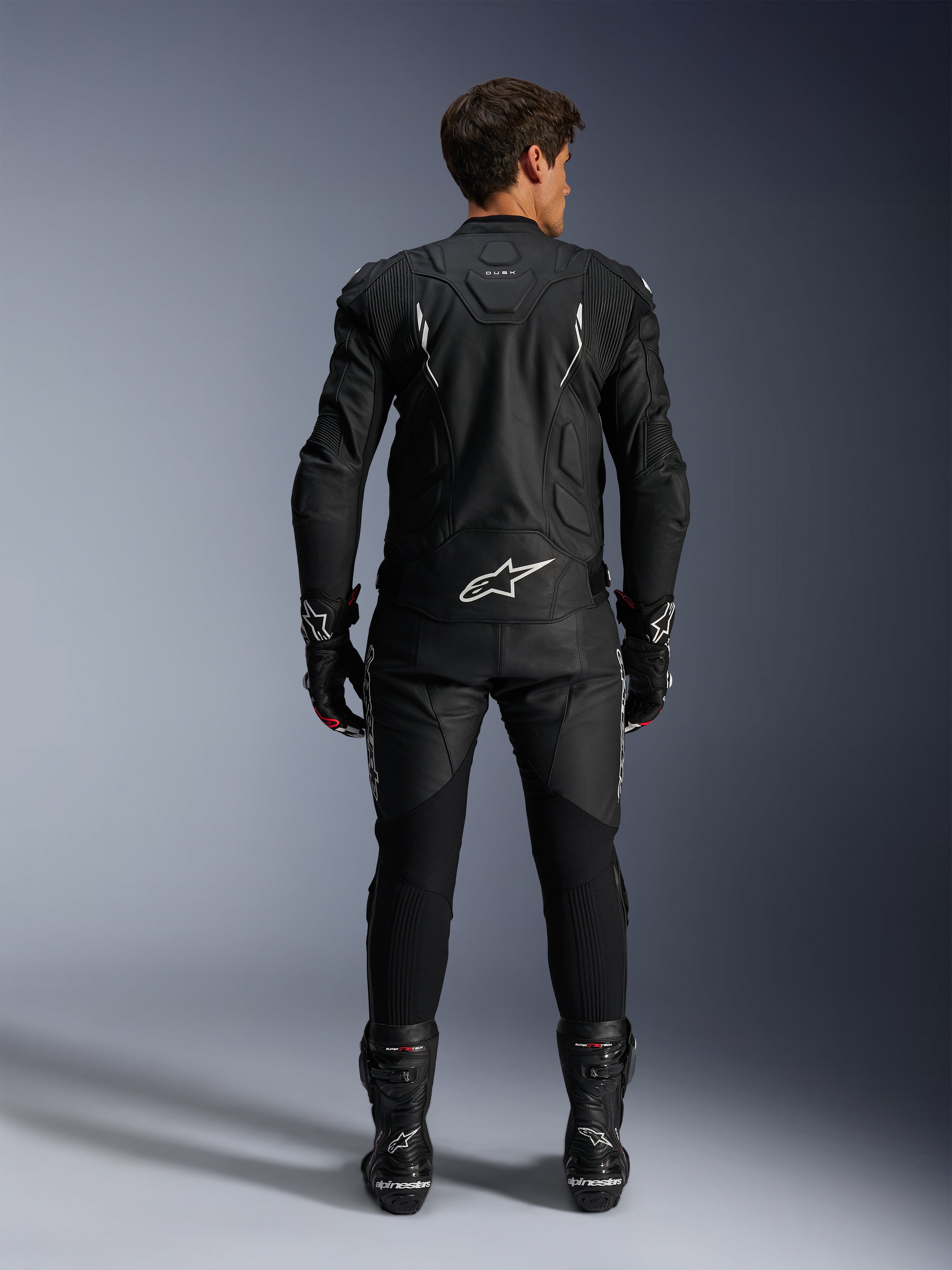 Pilota di moto in vista posteriore con giacca in pelle Alpinestars Dusk e pantaloni in pelle Missile V3, tuta in pelle nera con accenti bianchi, logo Alpinestars e marchio 'Dusk' sul retro, stivali e guanti racing neri, completo motorsport coordinato per corse su strada.