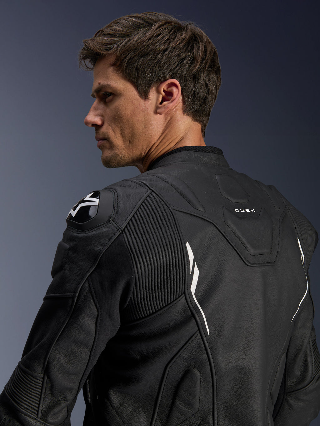 Giacca in pelle Alpinestars Dusk, nera con accenti bianchi, vista posteriore su modello, con protezioni per le spalle DFS e logo Alpinestars, pannelli a soffietto in pelle elasticizzata per la mobilità e marchio 'DUSK' sulla parte superiore della schiena, giacca da moto in pelle bovina premium per guida su strada e sportiva.