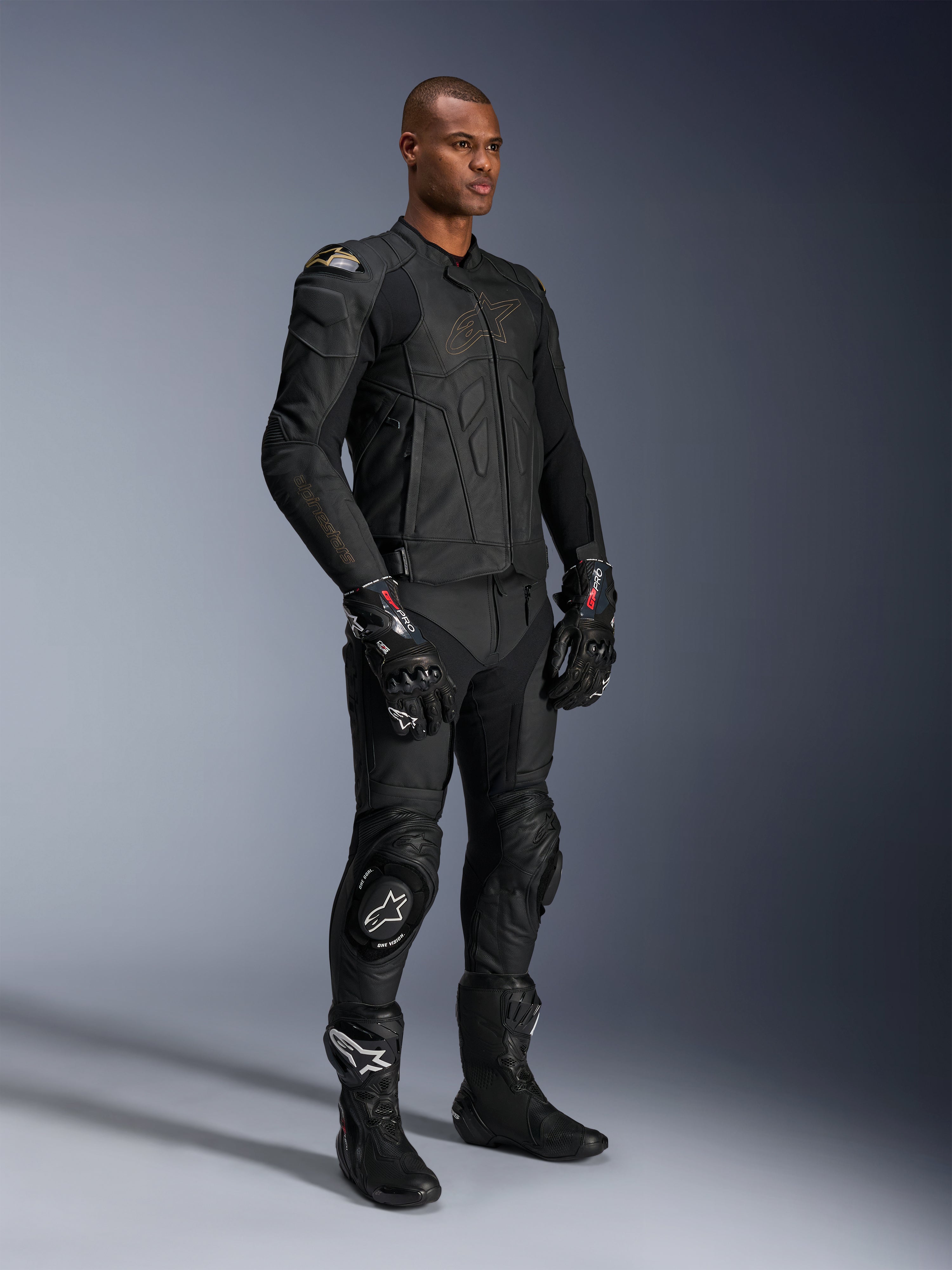 Modello che indossa pantaloni in pelle Alpinestars Missile V3 e giacca in pelle nera coordinata con dettagli oro, guanti racing GP Pro neri e stivali racing neri, equipaggiamento completo da moto per uso stradale, scatto in studio.