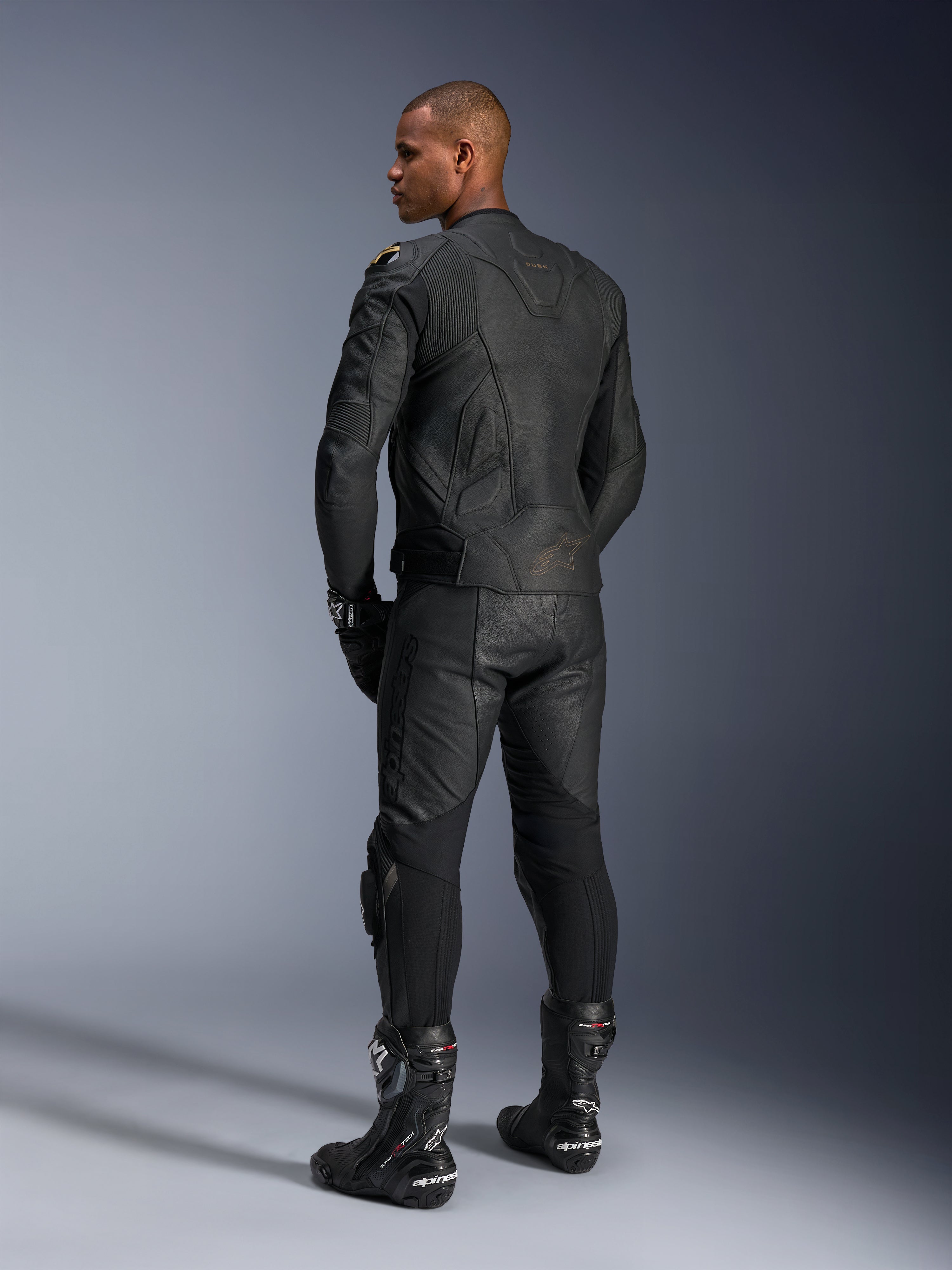 Pantaloni in pelle Alpinestars Missile V3 e giacca in pelle Dusk, nero con dettagli oro, vista posteriore del pilota, con loghi a rilievo e pannelli elasticizzati a soffietto, indossati con stivali e guanti Supertech R neri, abbigliamento professionale da motorsport.