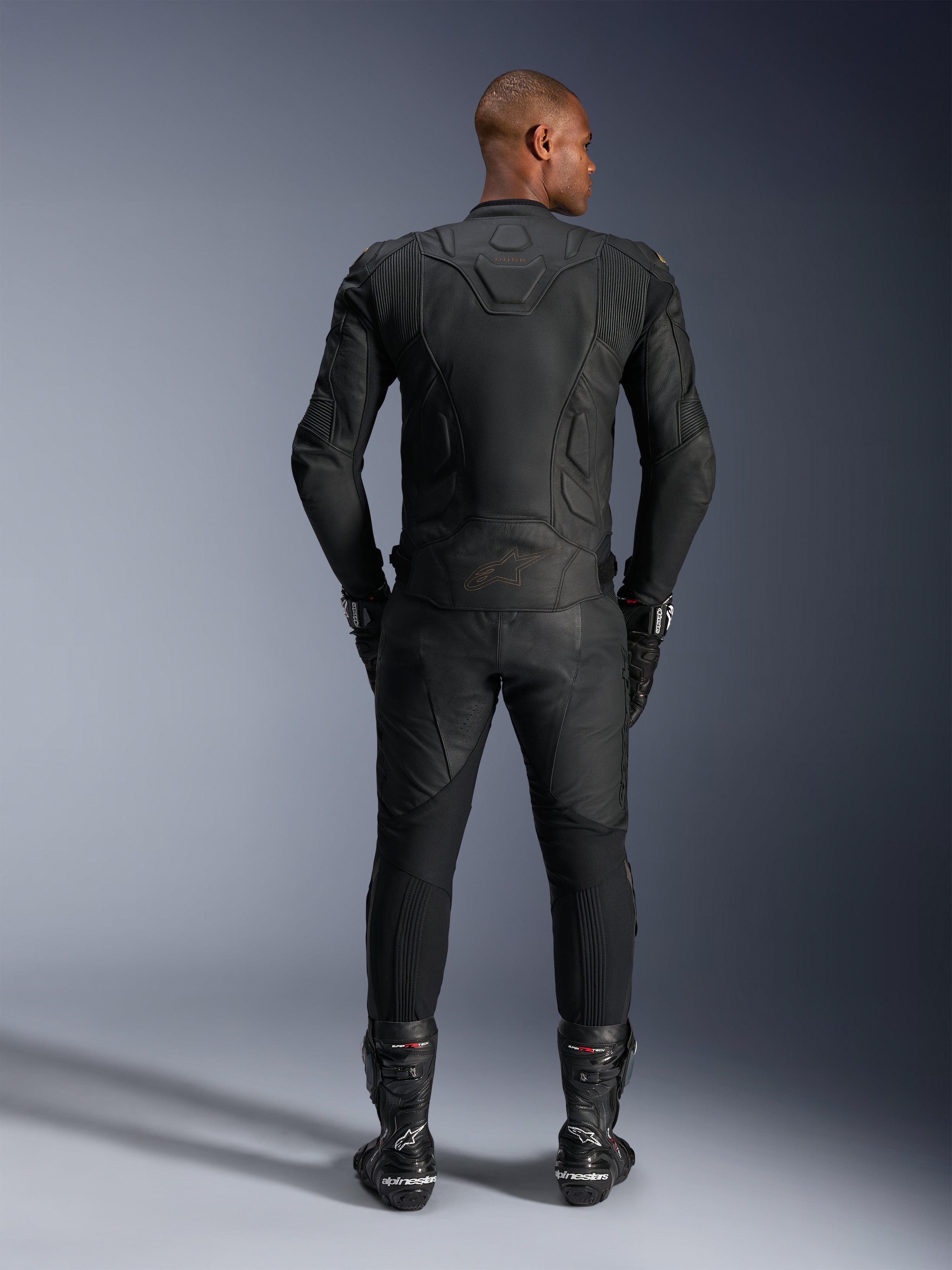 Alpinestars Dusk Leather Jacket, Black Laser, vista posteriore su un pilota, indossata con pantaloni da moto in pelle coordinati, guanti racing e stivali, con costruzione ergonomica multi-pannello 3D e pannelli protettivi in pelle.