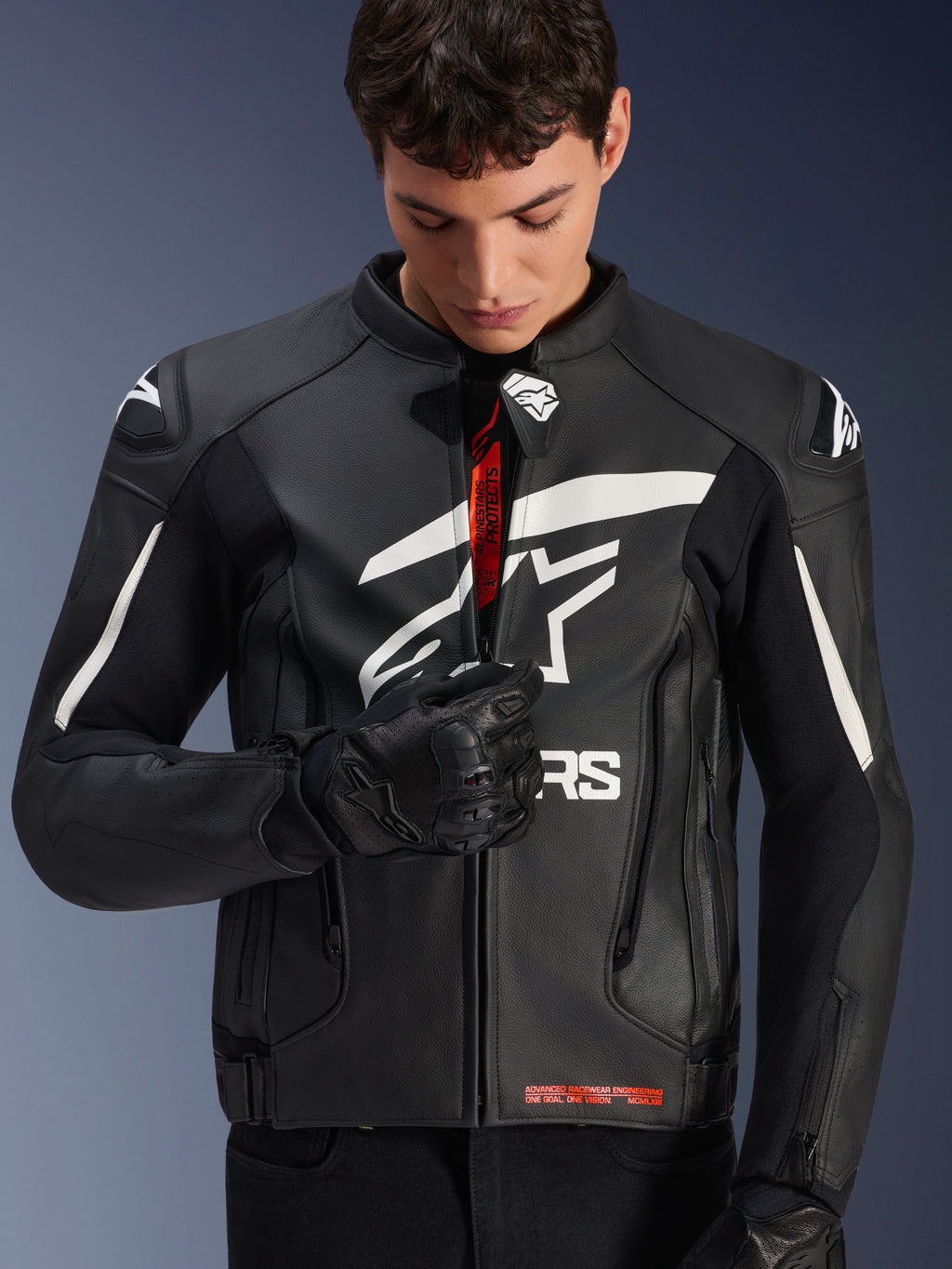 Giacca in pelle Alpinestars GP Plus V4, nera e bianca, modello maschile che chiude la zip frontale rivelando il marchio interno rosso, con evidenti loghi Alpinestars bianchi su petto e spalle, indossata con guanti da moto in pelle nera, abbigliamento da guida in pelle di derivazione sportiva