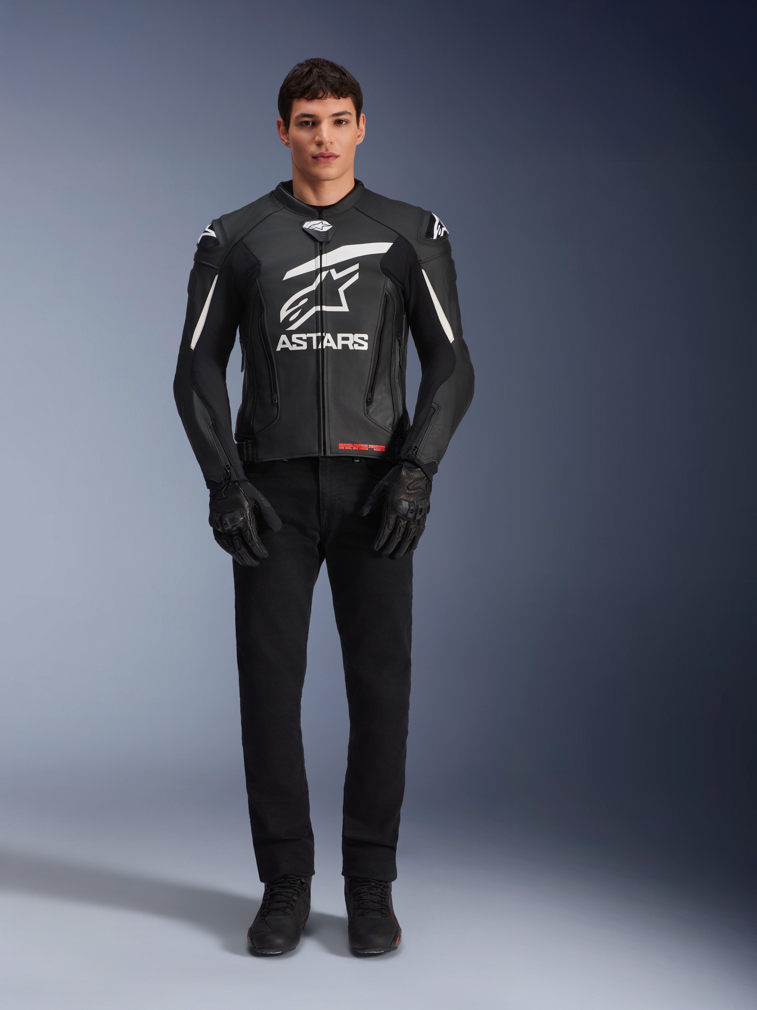 Modello che indossa la giacca in pelle Alpinestars GP Plus V4, nero e bianco, con vistoso marchio Astars bianco sul petto e dettagli bianchi sulle spalle, abbinata a guanti e pantaloni da moto neri, abbigliamento da moto sportivo ed elegante