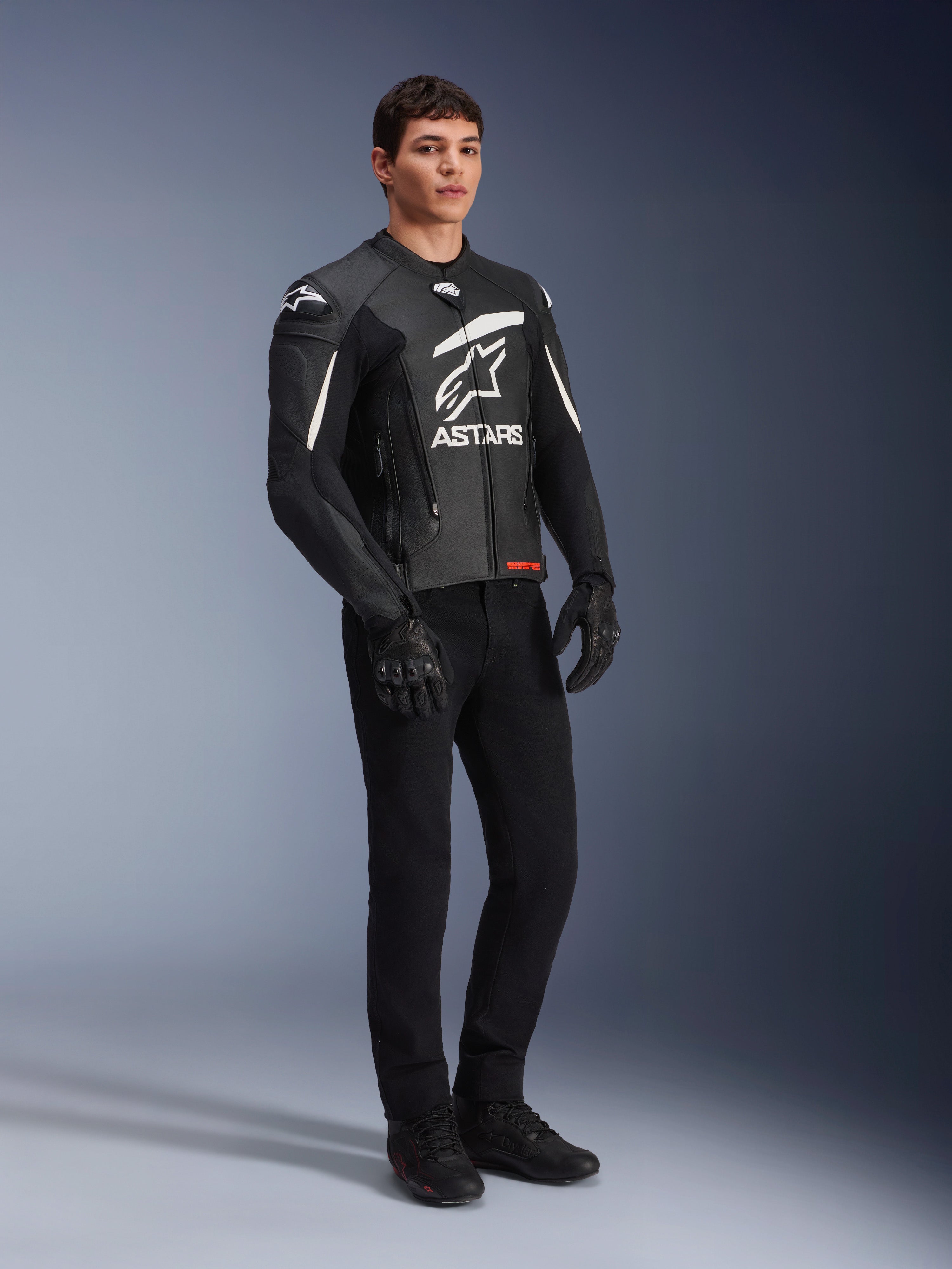 Giacca in pelle Alpinestars GP Plus V4, giacca da moto sportiva, nero e bianco, indossata da un modello uomo con pantaloni da moto e guanti neri, con un grande logo ASTARS bianco sul petto e slider esterni DFS sulle spalle, design professionale di derivazione racing