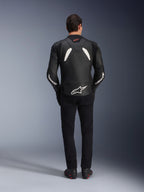 Modello visto da dietro che indossa la giacca in pelle Alpinestars GP Plus V4 in nero e bianco, abbinata a pantaloni neri e stivali da moto, con pannelli bianchi sulle spalle e logo Alpinestars sulle maniche e sulla parte bassa della schiena.