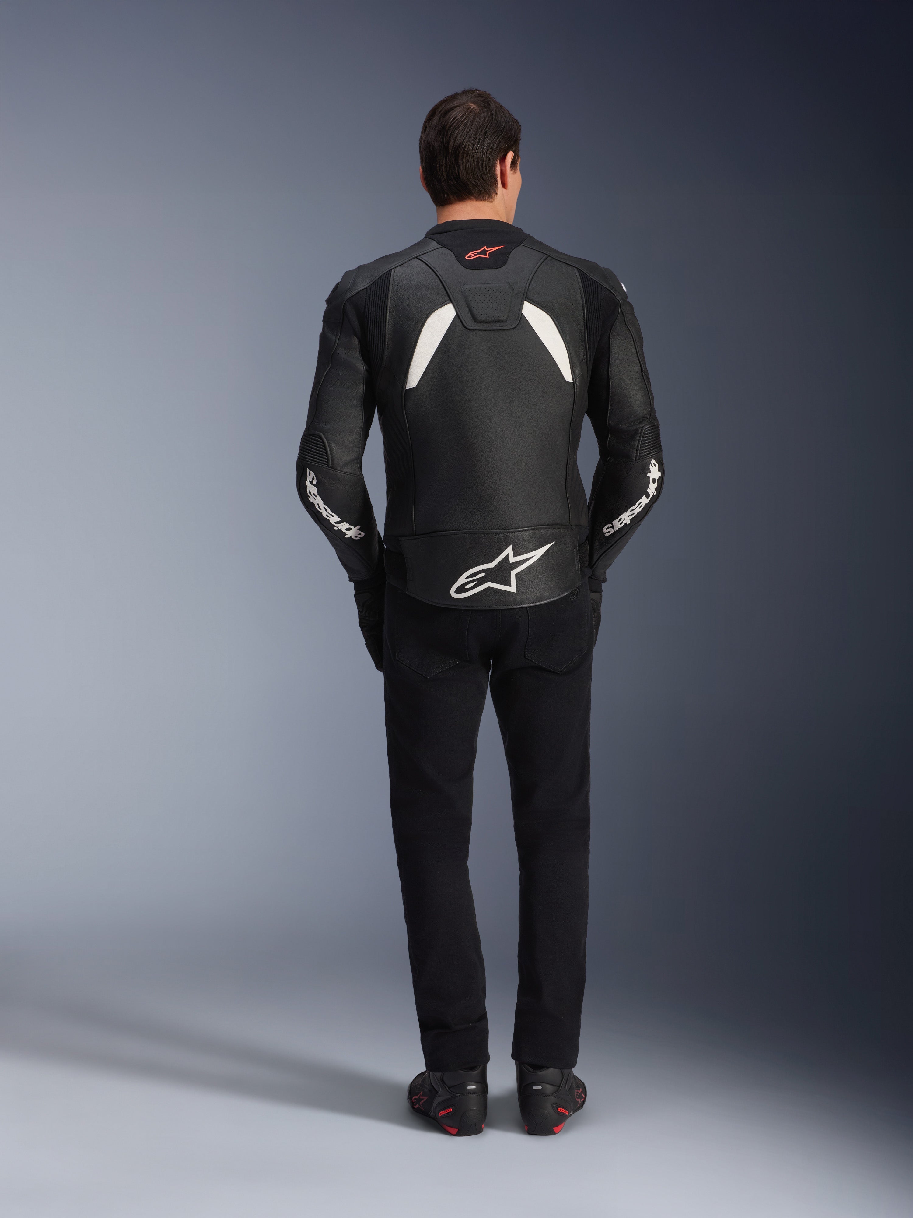 Modello visto da dietro che indossa la giacca in pelle Alpinestars GP Plus V4 in nero e bianco, abbinata a pantaloni neri e stivali da moto, con pannelli bianchi sulle spalle e logo Alpinestars sulle maniche e sulla parte bassa della schiena.
