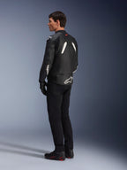 Giacca in pelle Alpinestars GP Plus V4, giacca sportiva in pelle, nero e bianco, indossata da un modello in una vista di profilo posteriore-laterale, caratterizzata da loghi Alpinestars bianchi sulla parte bassa della schiena e sulla manica, abbinata a pantaloni da moto neri e stivali tecnici per un look sportivo performante.