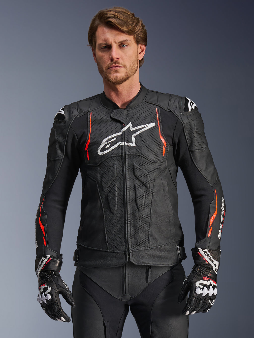 Alpinestars Dusk Airflow Leather Jacket, giacca racing in pelle nera con accenti rosso fluo e bianchi, dotata di ampie perforazioni per la ventilazione, logo bianco sul petto e slider esterni sulle spalle, indossata da un modello con guanti racing coordinati