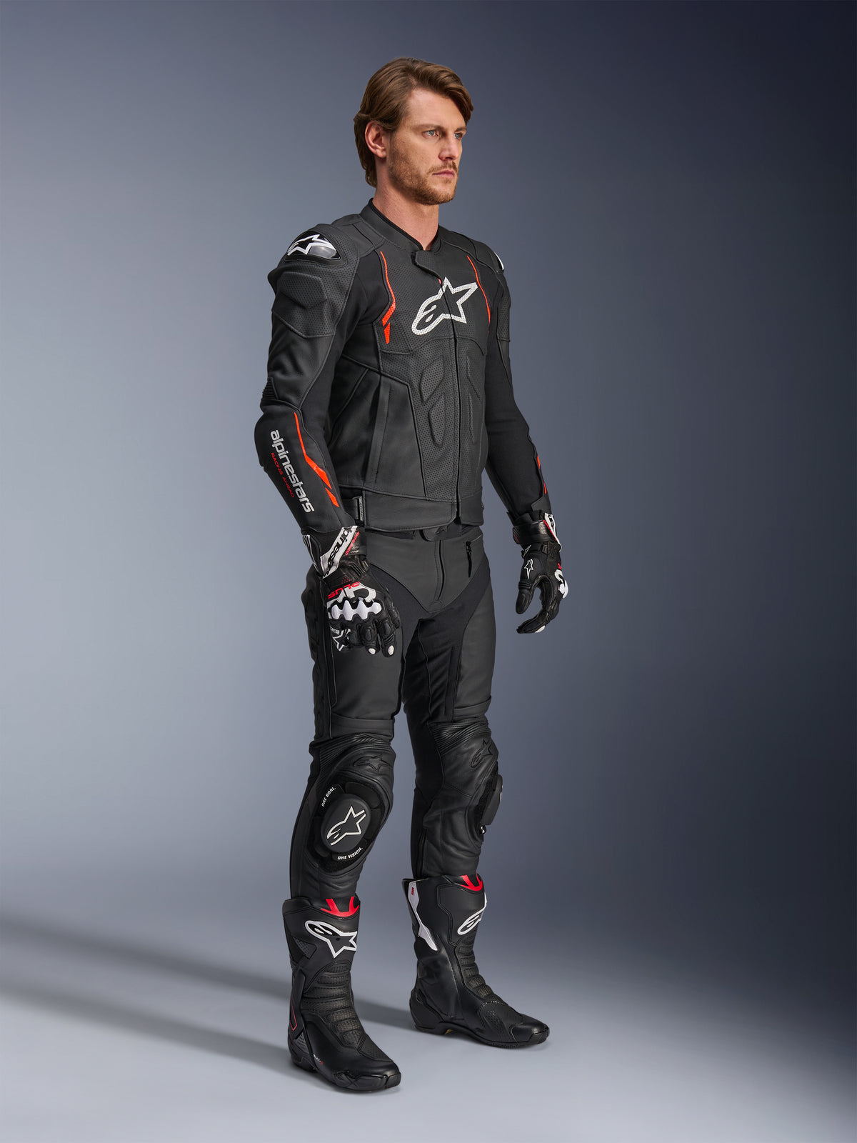 Giacca in pelle Alpinestars Dusk Airflow, Nero Rosso Fluo, indossata come parte di un outfit da corsa completo con pantaloni in pelle, guanti e stivali coordinati, caratterizzata da pelle traforata e accenti sportivi.