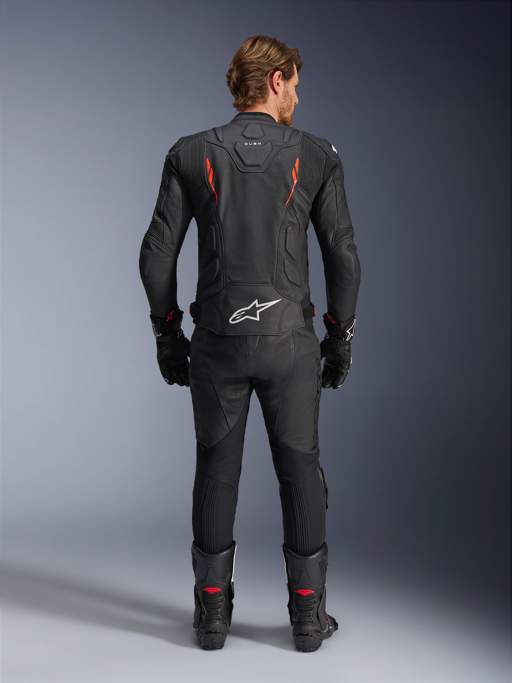 Giacca in pelle Alpinestars Dusk Airflow, nero e rosso fluo, vista posteriore che mostra le ampie perforazioni e il grande logo bianco sulla parte bassa della schiena, indossata con abbigliamento racing in pelle coordinato, inclusi pantaloni, stivali e guanti