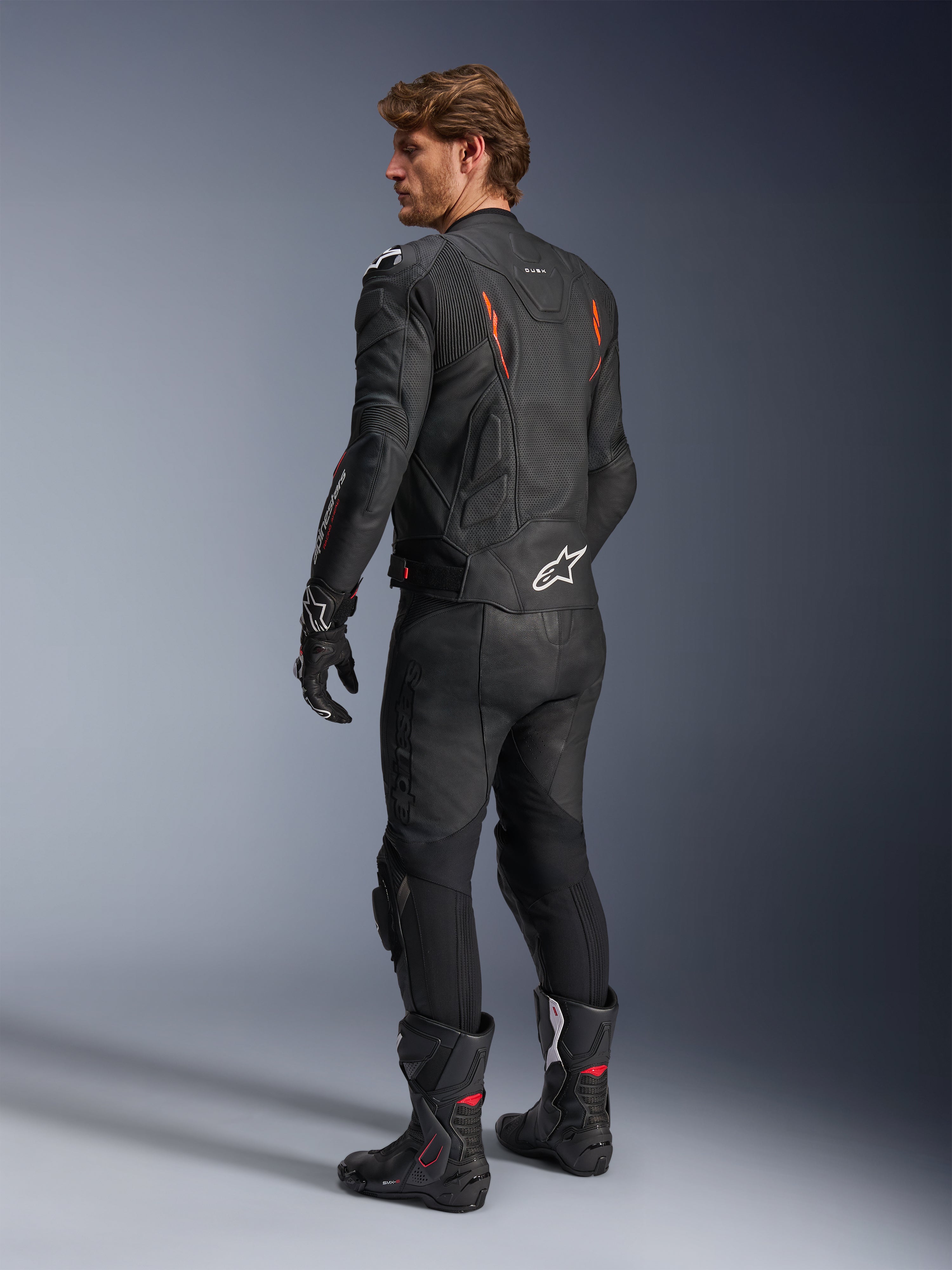 Giacca in pelle Alpinestars Dusk Airflow, nero con accenti rosso fluo, vista posteriore di un pilota con abbigliamento da moto completo in pelle, inclusi pantaloni neri coordinati, guanti e stivali racing, caratterizzata da pelle traforata e loghi Alpinestars bianchi.
