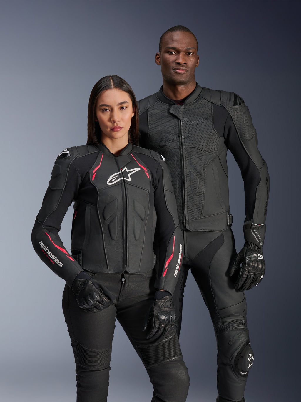 Giacca in pelle Alpinestars Stella Dusk, Nero Diva Pink Bianco, indossata da una modella accanto a un modello in abbigliamento in pelle nera coordinato, con accenti rosa e bianchi, logo a stella bianco sul petto e guanti da moto neri