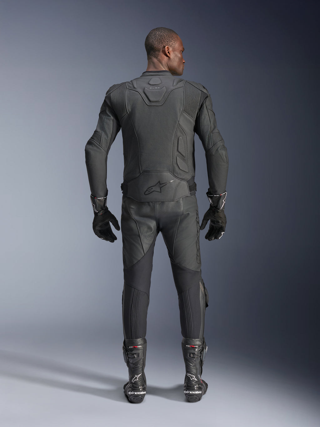 Modello uomo visto da dietro che indossa la giacca in pelle Alpinestars Dusk Airflow in nero, abbinata a pantaloni da corsa in pelle, guanti e stivali neri coordinati, con ampie perforazioni per la ventilazione e il logo Alpinestars sulla parte bassa della schiena.