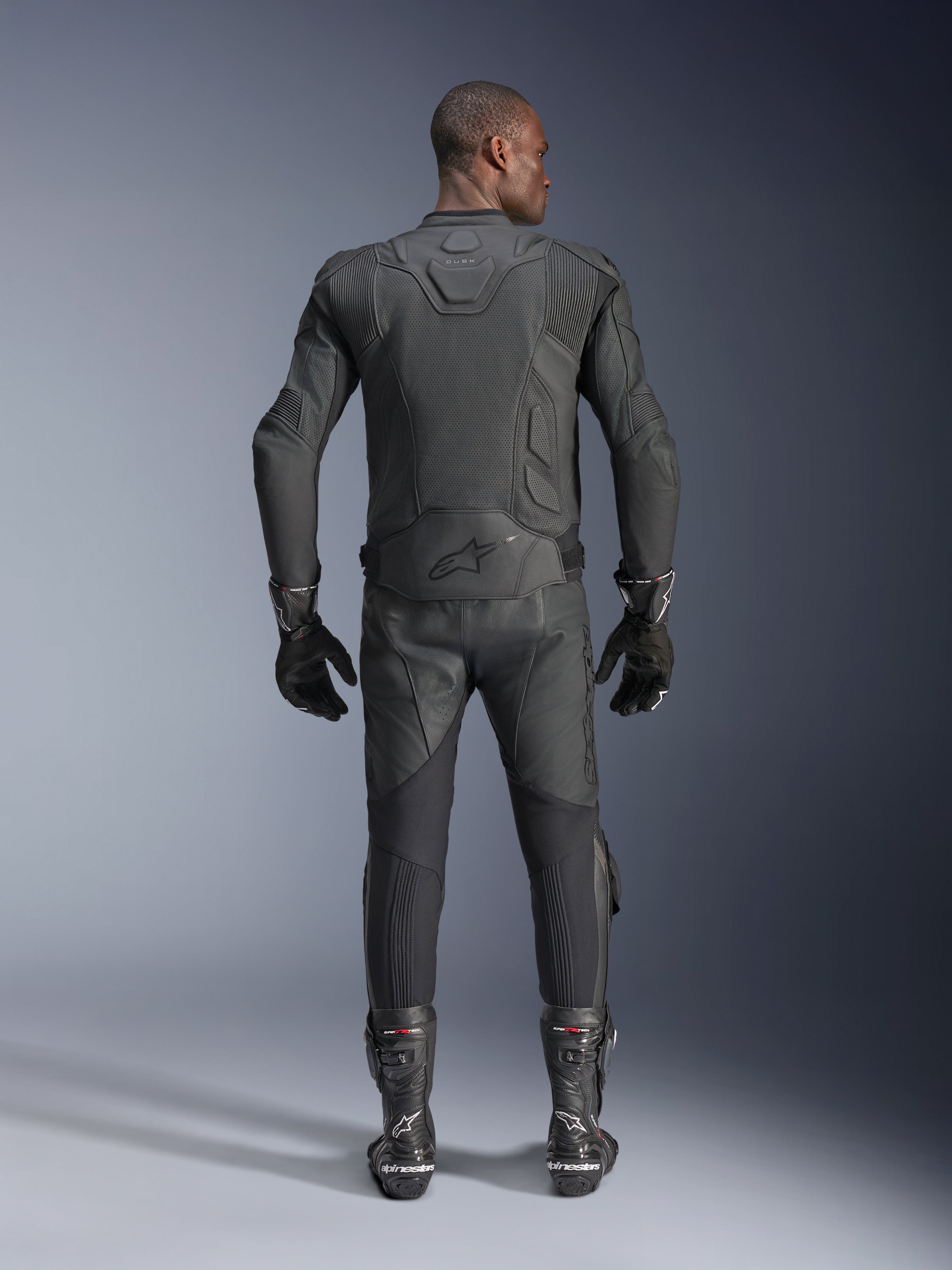Modello uomo visto da dietro che indossa la giacca in pelle Alpinestars Dusk Airflow in nero, abbinata a pantaloni da corsa in pelle, guanti e stivali neri coordinati, con ampie perforazioni per la ventilazione e il logo Alpinestars sulla parte bassa della schiena.