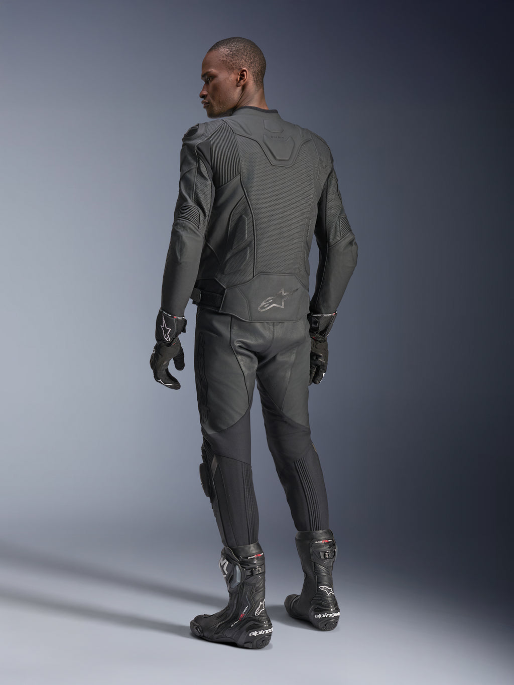 Pilota uomo visto da dietro che indossa la giacca in pelle Alpinestars Dusk Airflow in Black Black, abbinata a pantaloni, guanti e stivali in pelle coordinati, mettendo in risalto le ampie perforazioni sulla schiena e la vestibilità sportiva.